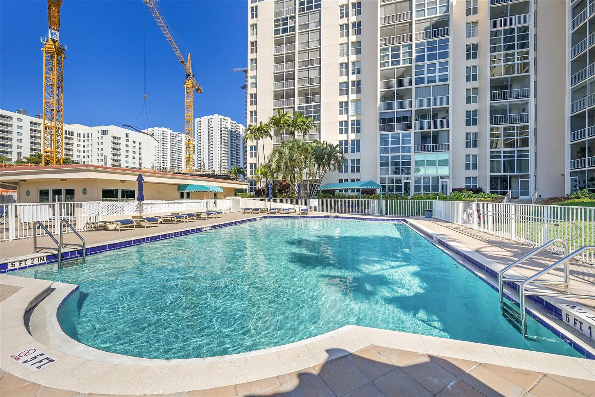 Outdoor pool beside a high-rise condominium, with lounge chairs, palm trees, and construction cranes nearby.