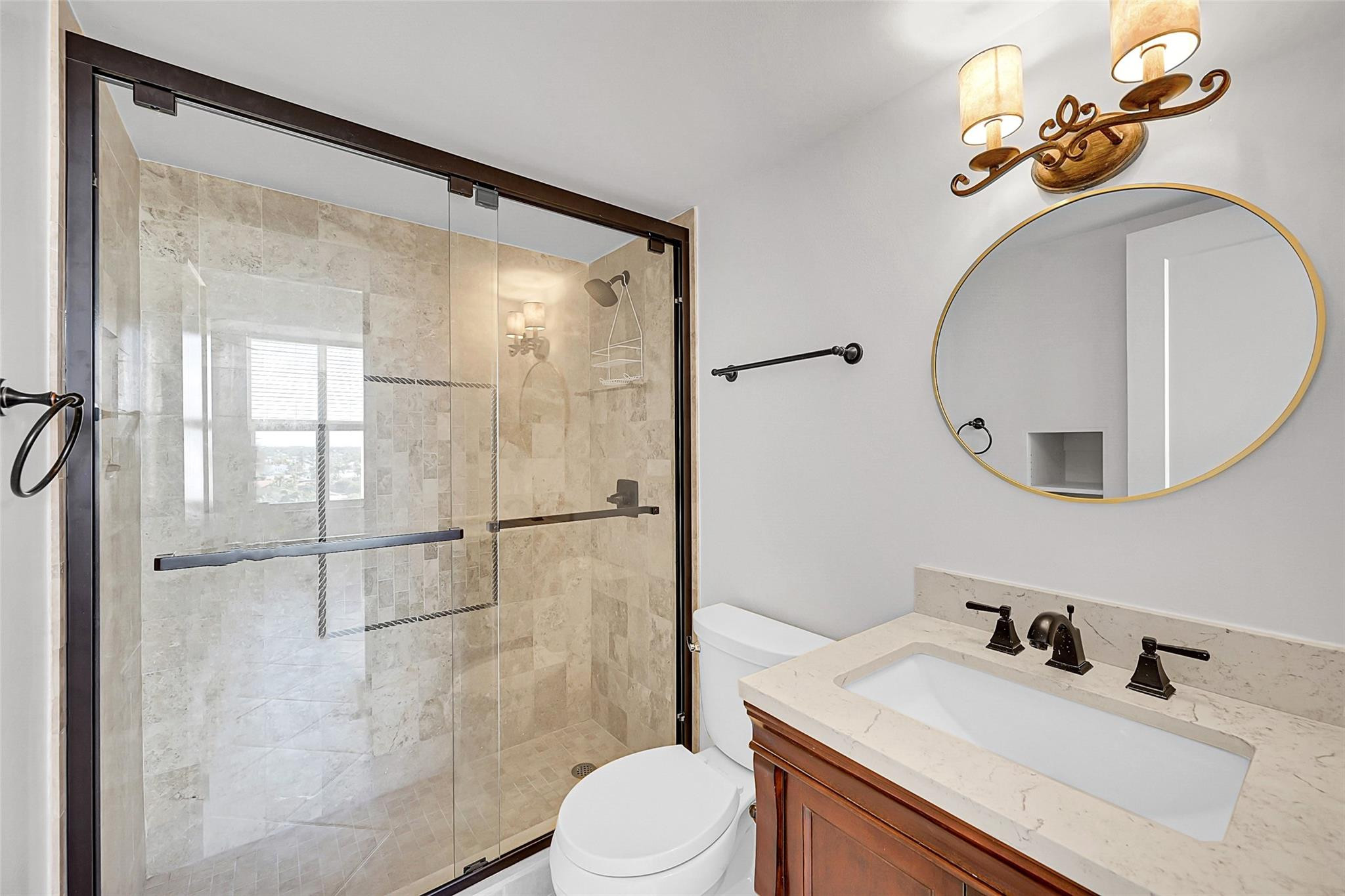 Bathroom with a glass-enclosed stone-tile shower and wood vanity beneath a round gold-rimmed mirror.