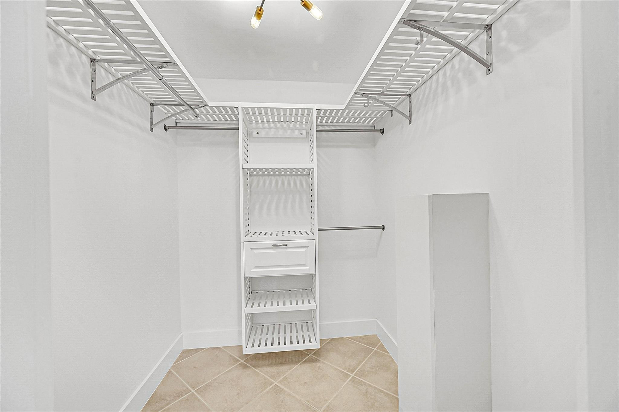 Walk-in closet with white shelving, hanging rods, a central drawer, and beige tile flooring.