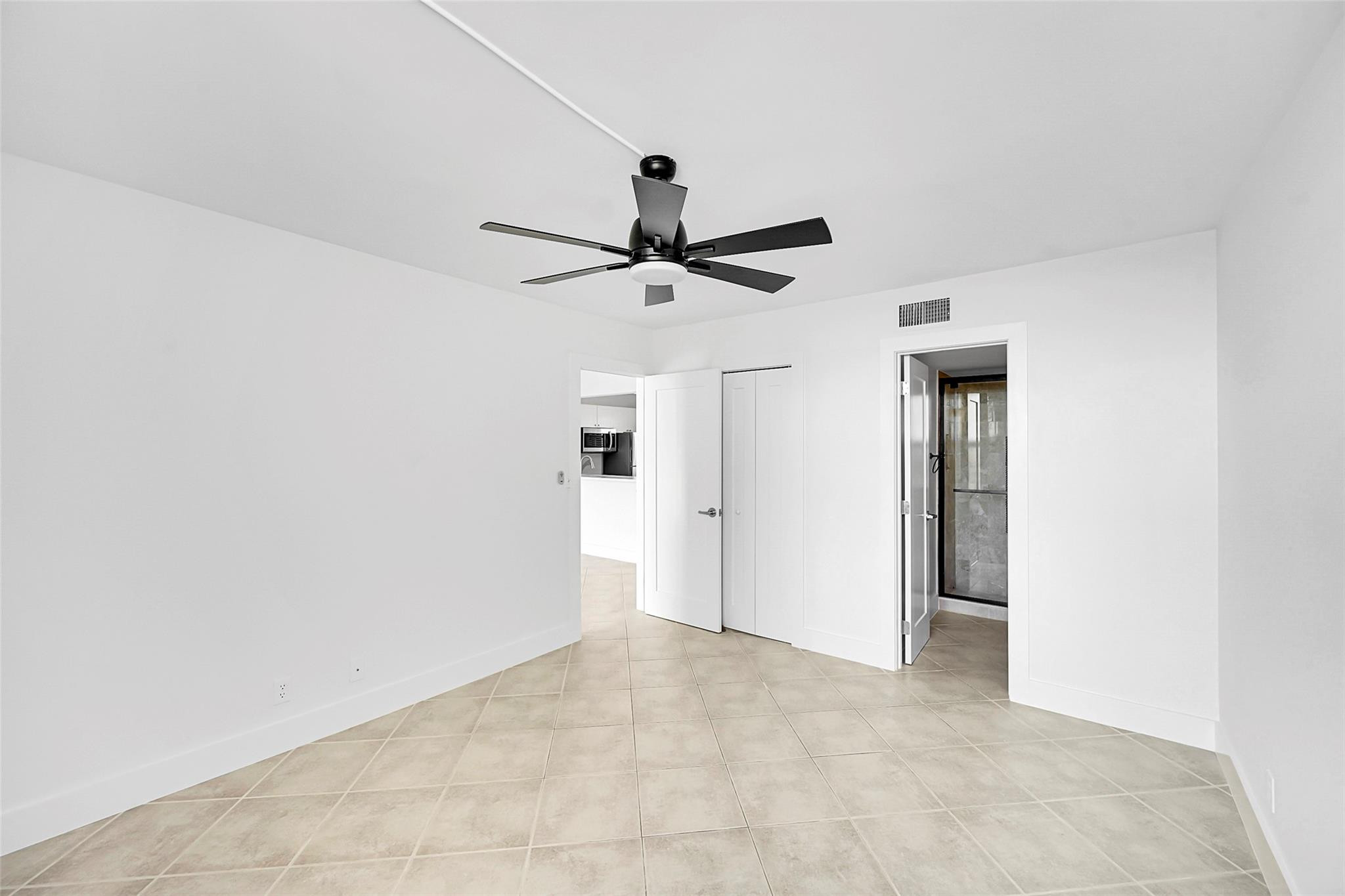 Empty bedroom with beige tile floors, a black ceiling fan, and open doors to adjoining rooms.