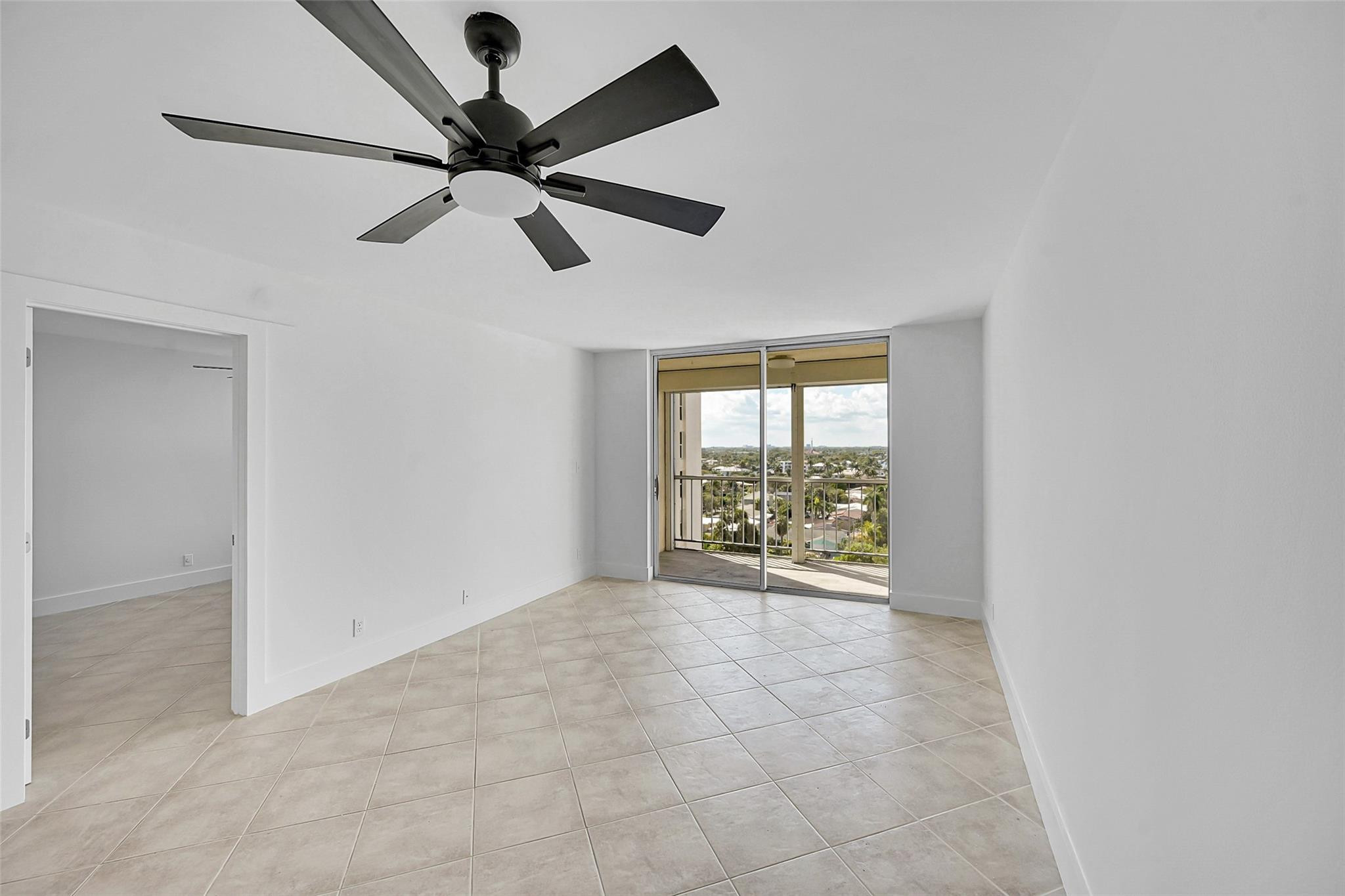 Empty tiled living room with a black ceiling fan and sliding doors to a neighborhood-view balcony.