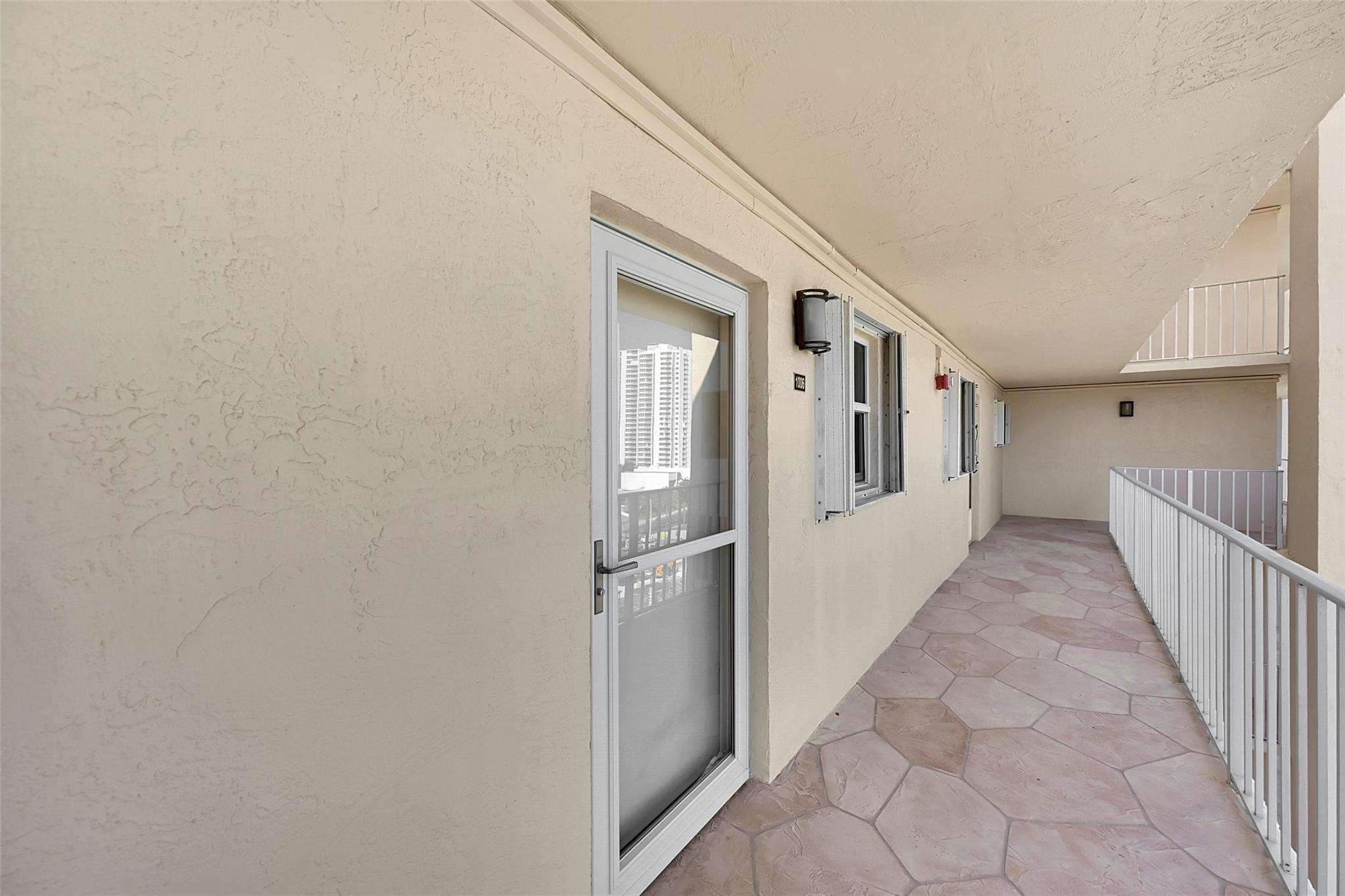 Covered condominium corridor with a screened entrance door, white railings, and hexagonal tile flooring.