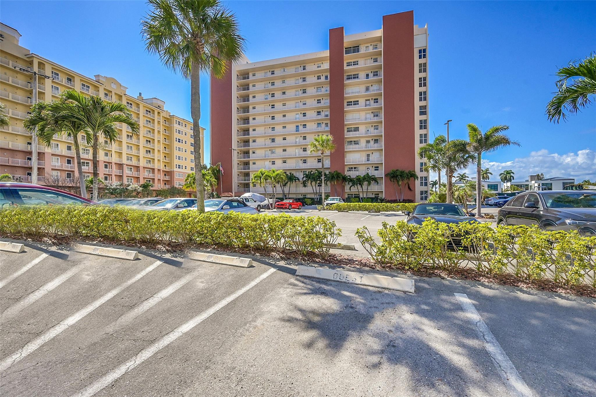 Condominium exterior with red facade sections, balconies, palm trees, and a parking lot.