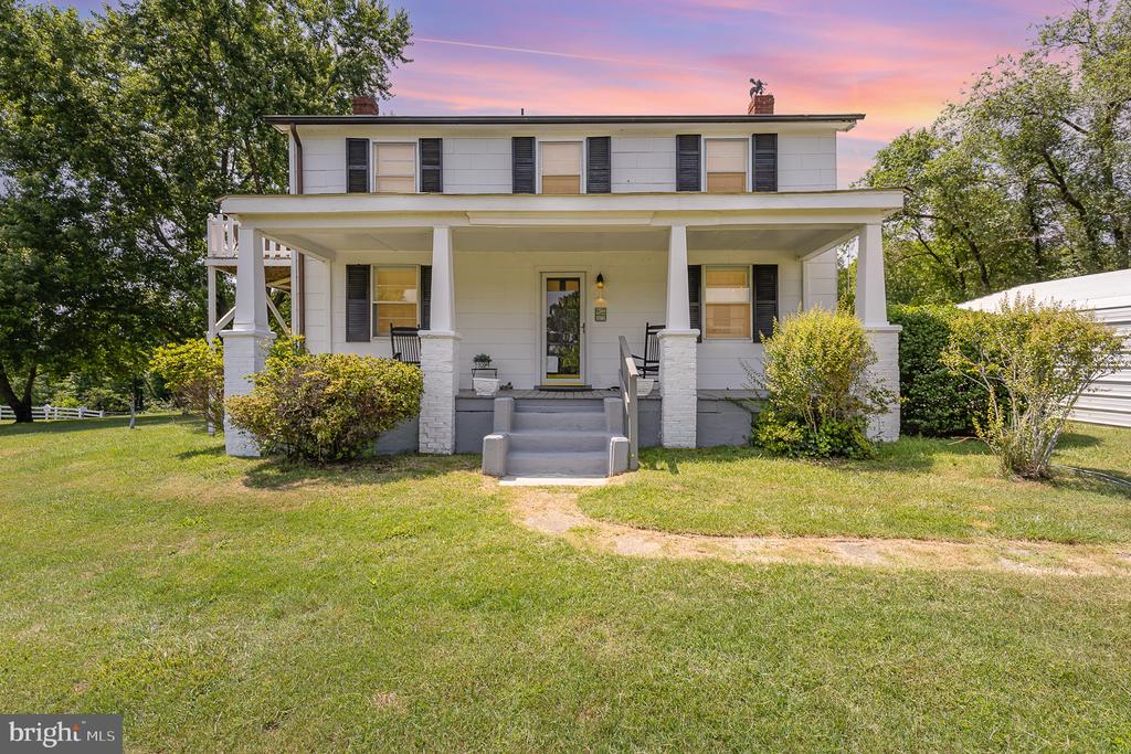 White two-story house with a broad covered porch, dark shutters, and a front lawn at sunset.