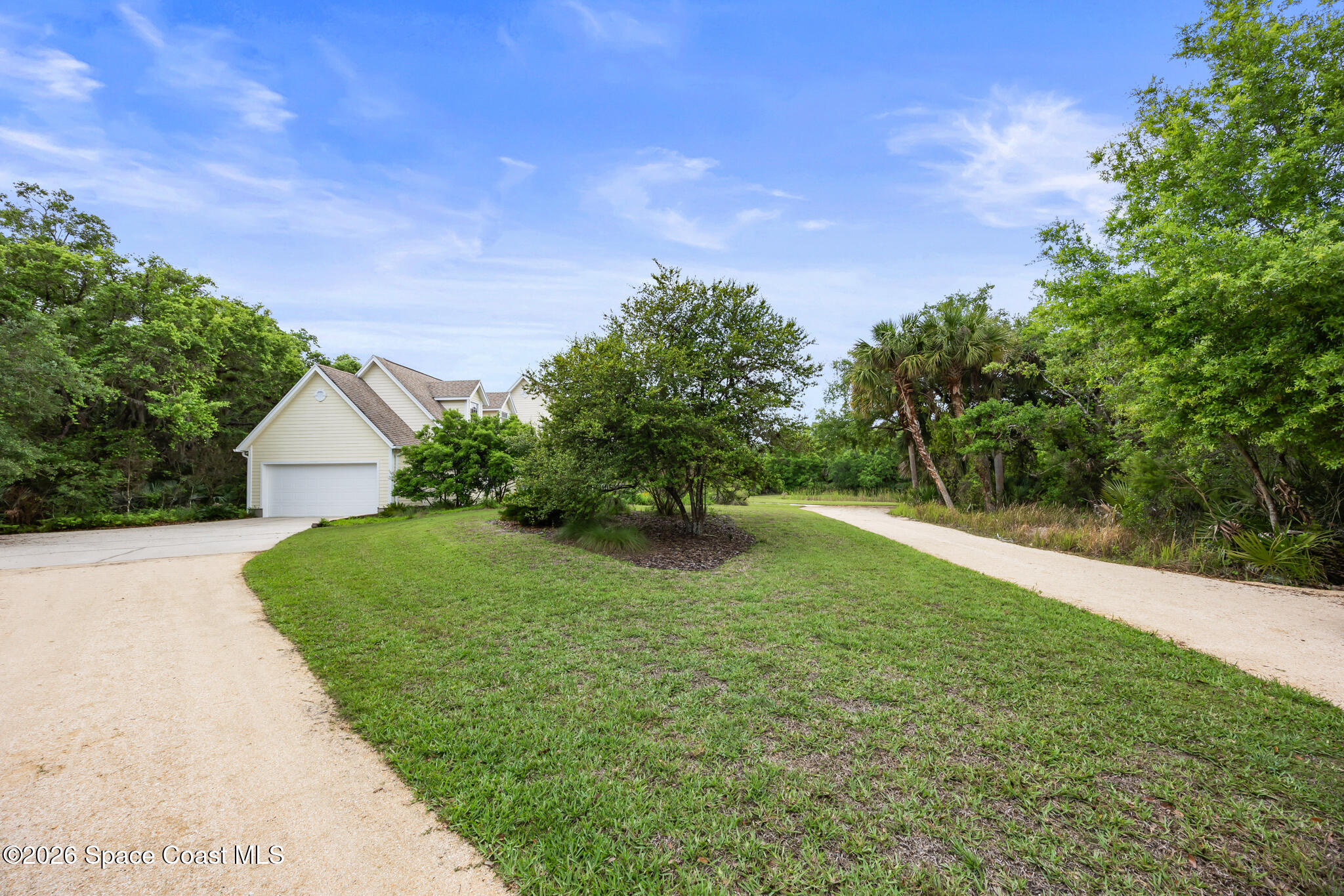 House with a white garage, curved gravel driveway, and wide lawn surrounded by trees.