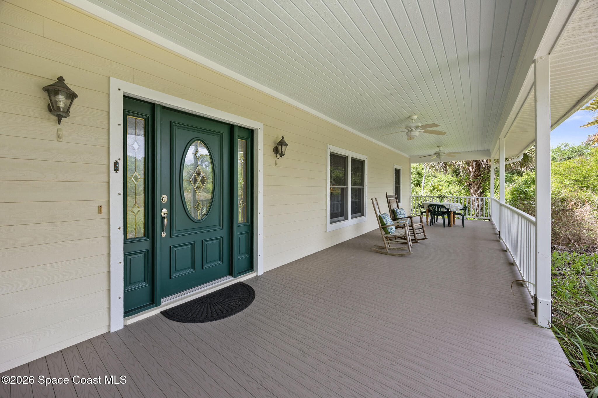 Covered front porch with a dark green glass-paneled door, rocking chairs, and a table beside tropical greenery.