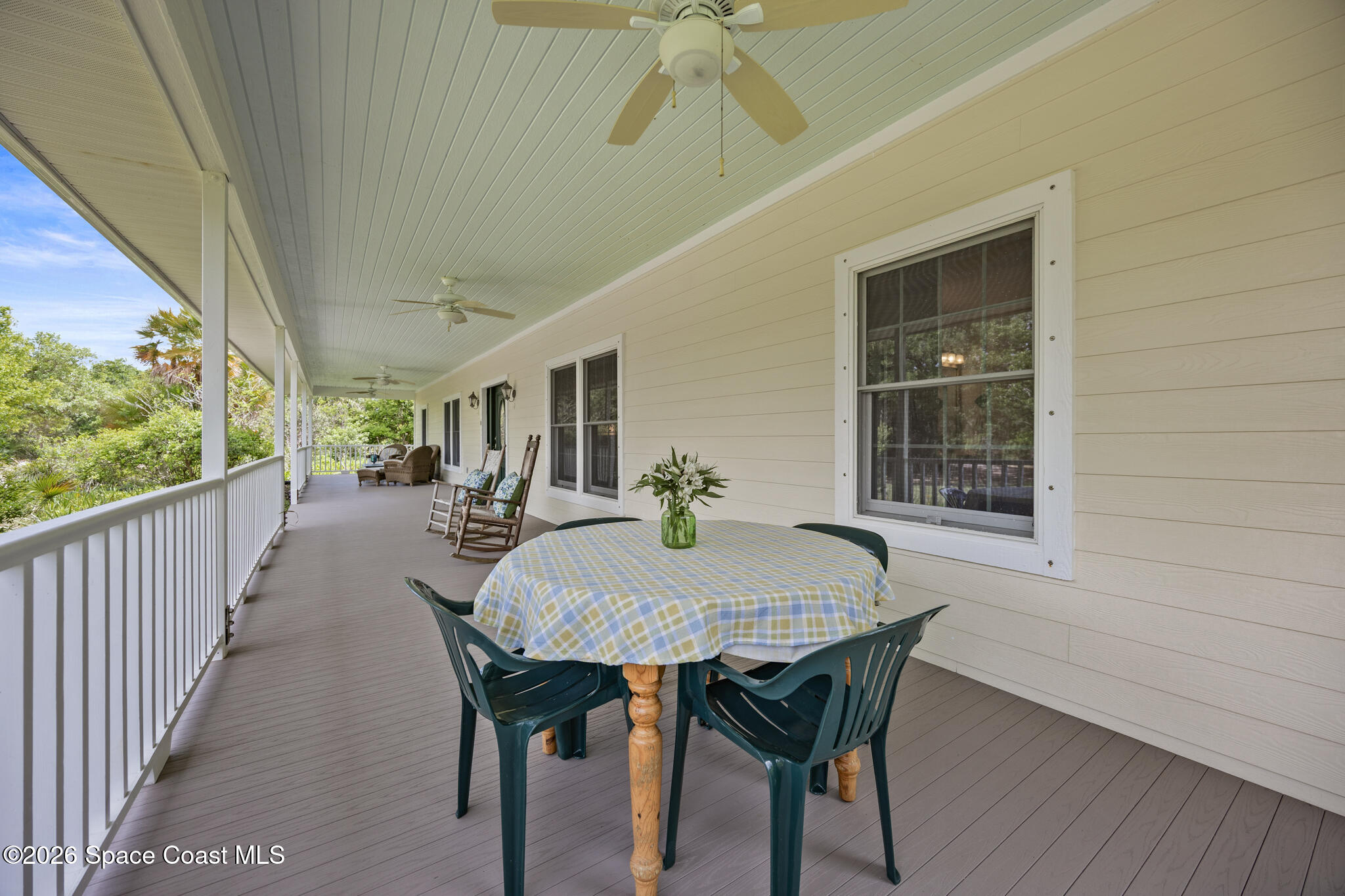 Covered porch with ceiling fans, rocking chairs, and a round dining table beside leafy trees.