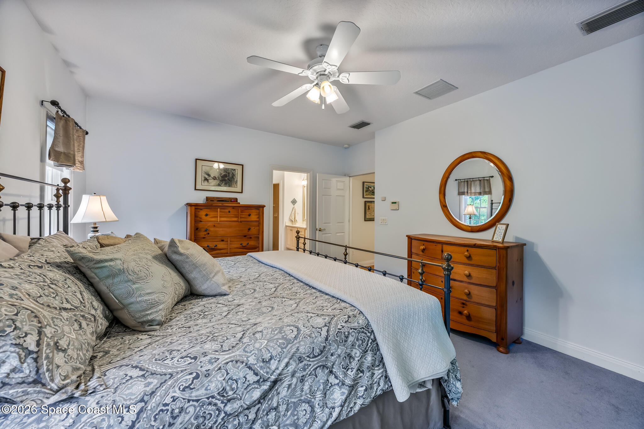 Furnished bedroom with paisley bedding, wooden dressers, and a large round mirror.