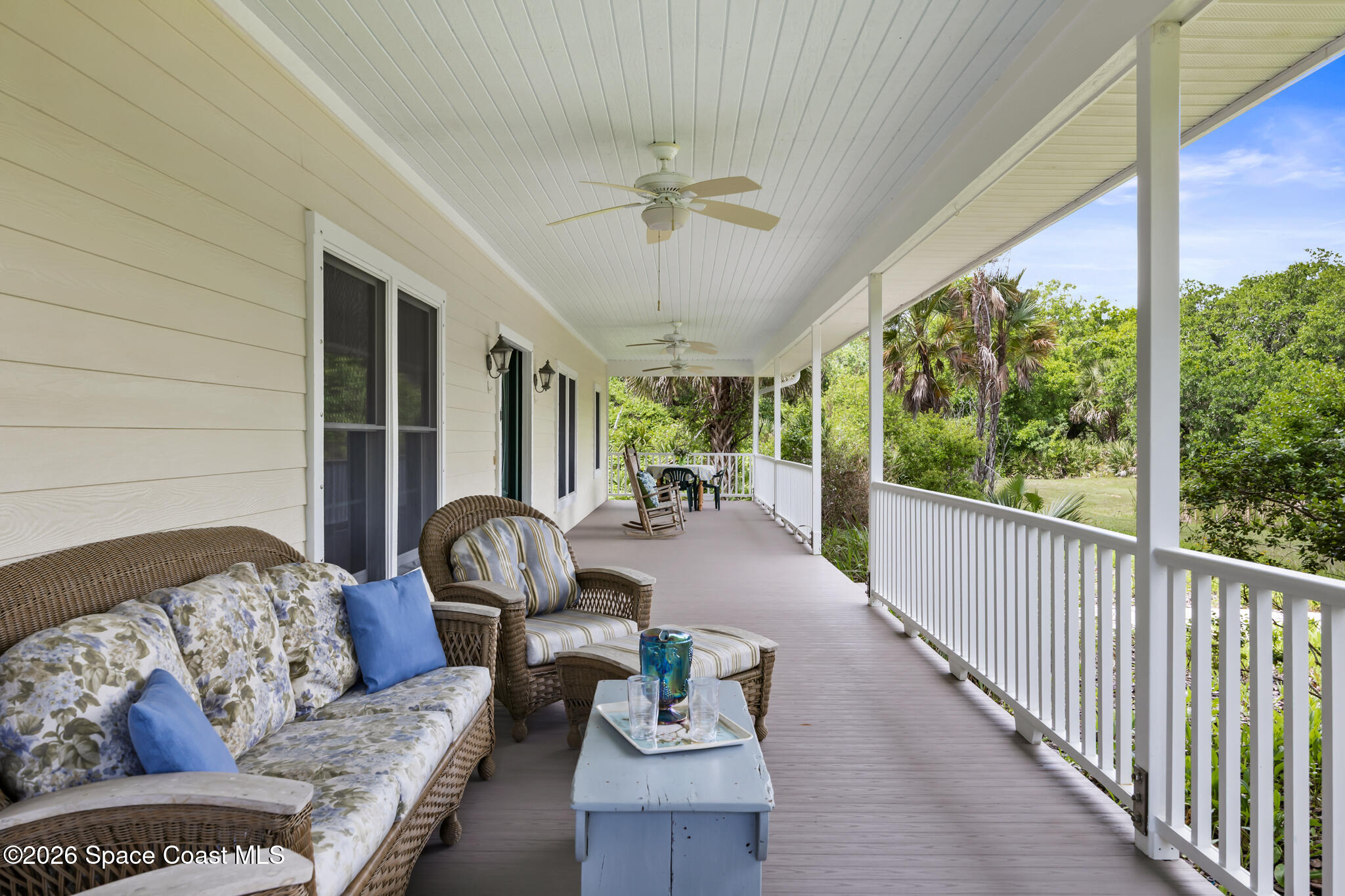 Covered porch with wicker seating and ceiling fans overlooking a green, palm-filled yard.
