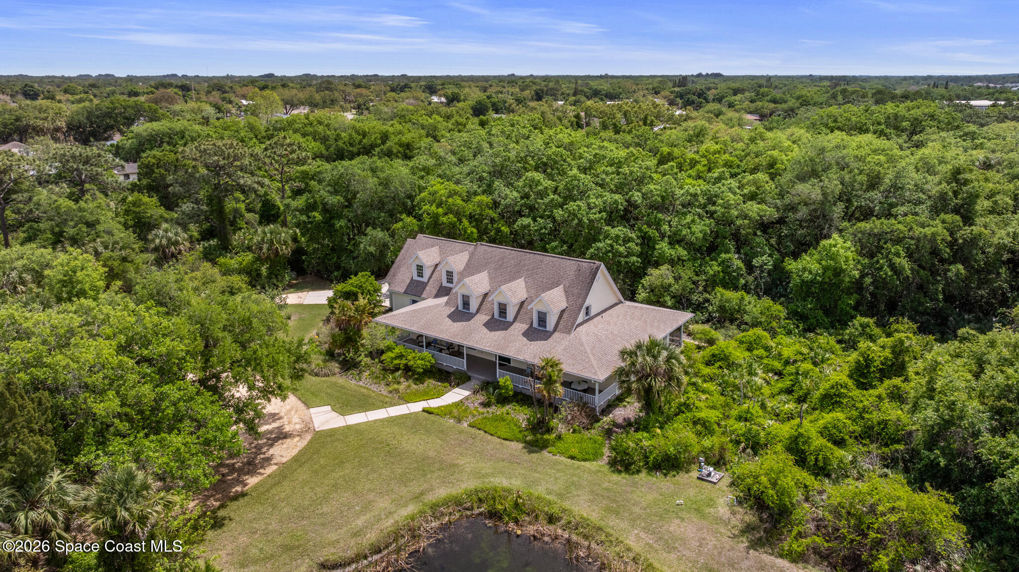 Aerial view of a large gabled house surrounded by trees, with a lawn, small pond, and wraparound porch.