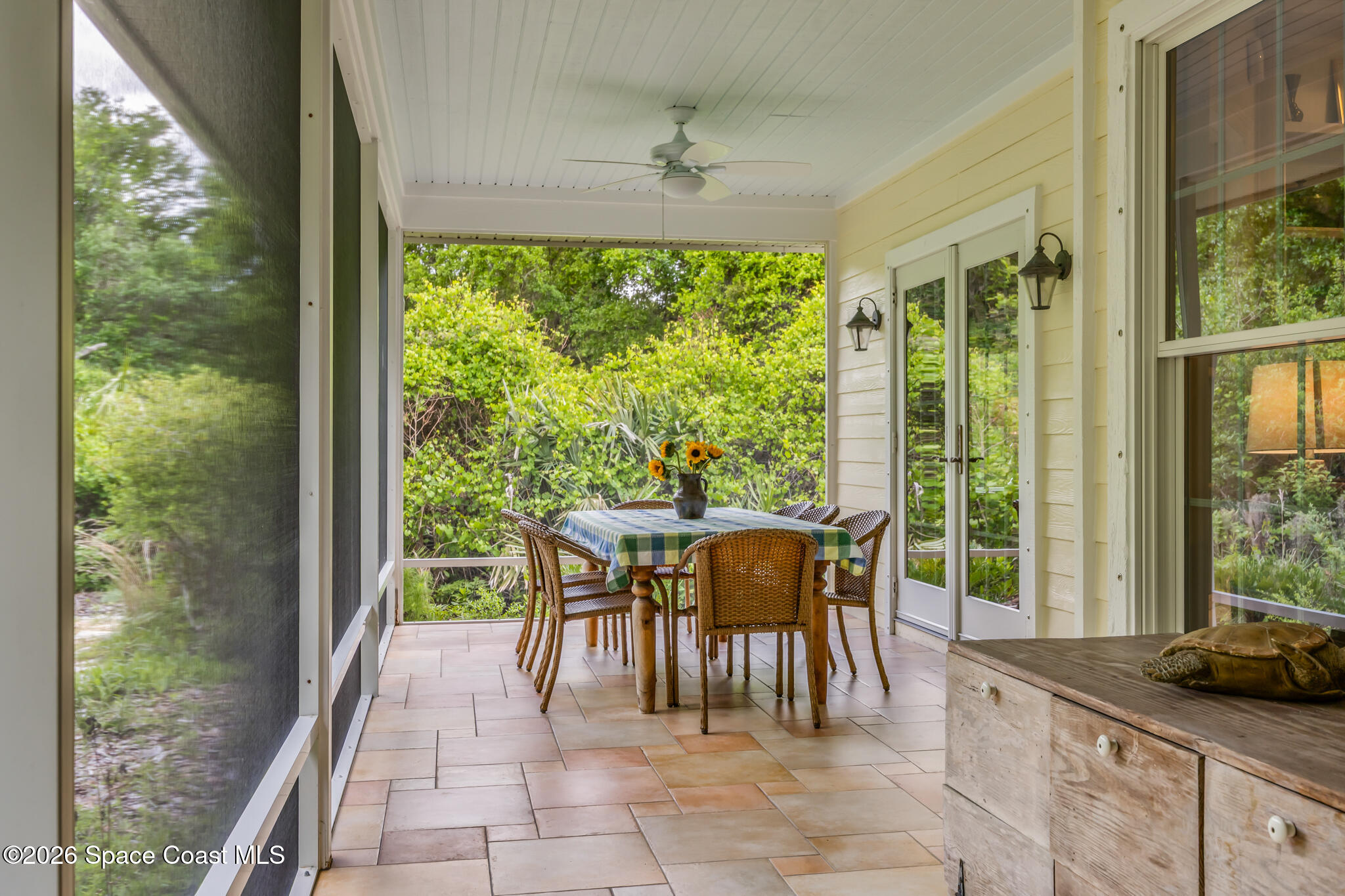 Screened porch with wicker dining set, tiled floor, and dense garden greenery.