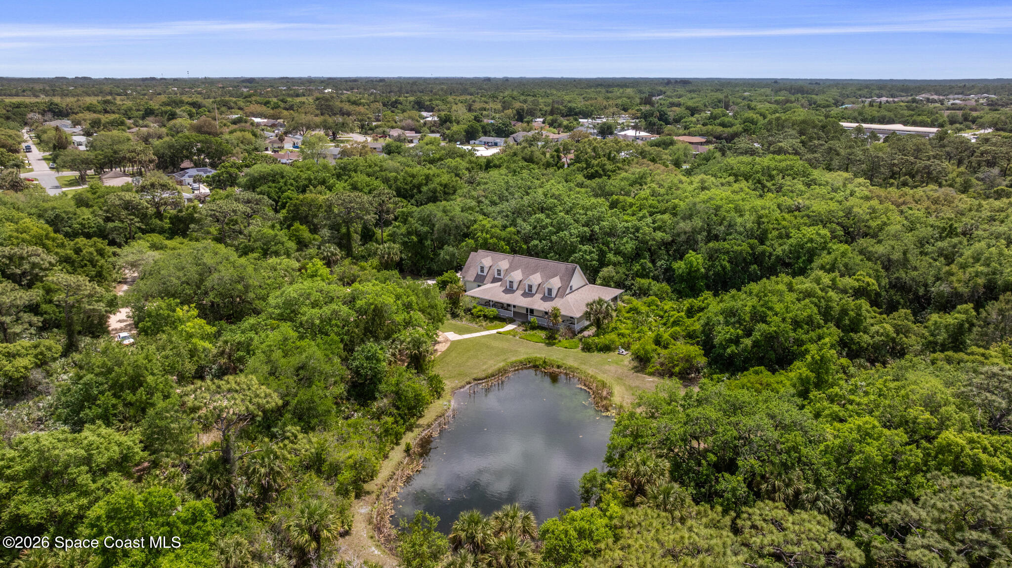 Aerial view of a house amid dense trees, with a pond in front and homes in the distance.