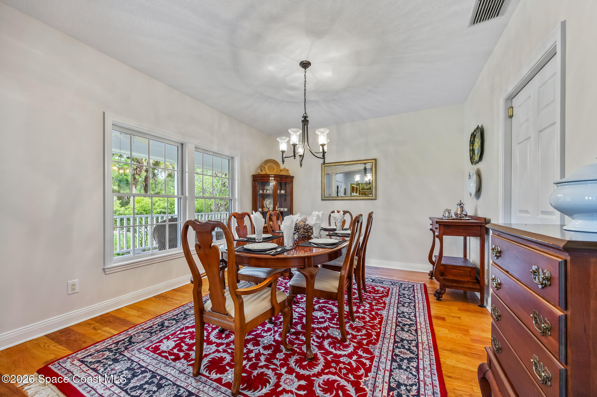 Dining room with a polished wooden table, red patterned rug, chandelier, and windows overlooking greenery.