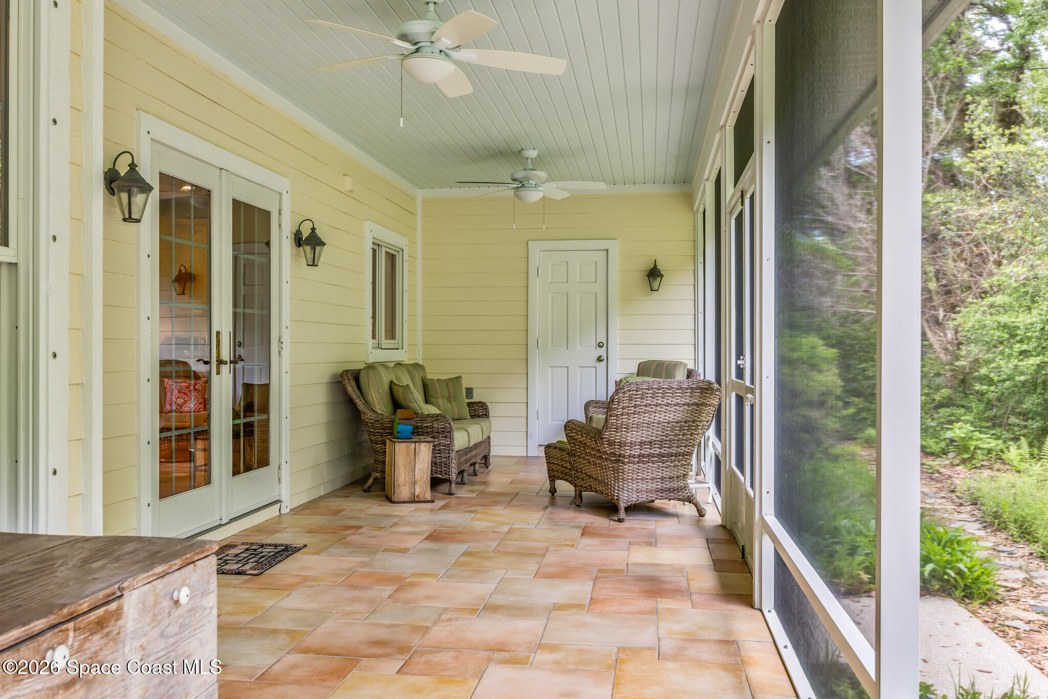 Screened patio with wicker seating, tile flooring, and ceiling fans beside a garden.