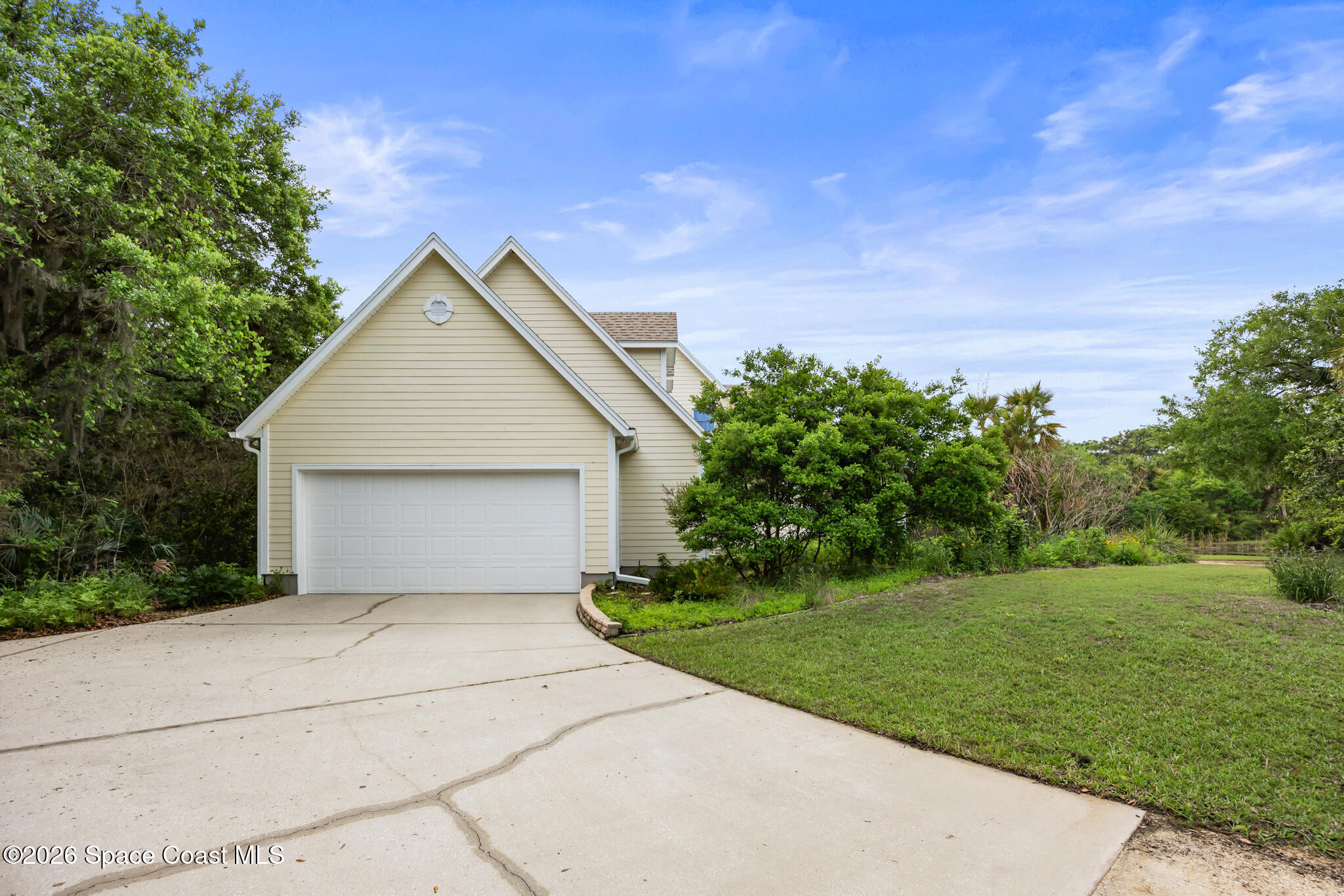 Beige-sided home with a white double garage, wide concrete driveway, and mature greenery.
