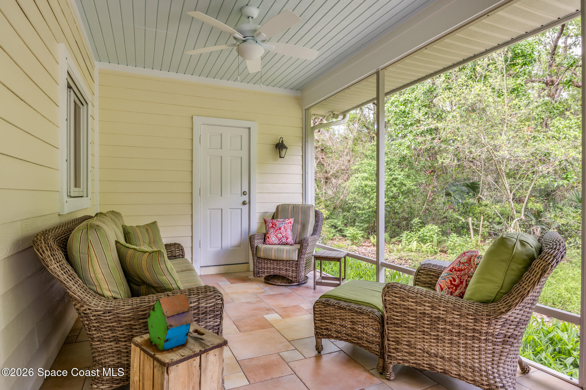 Screened-in porch with wicker seating and a ceiling fan overlooking a lush wooded yard.