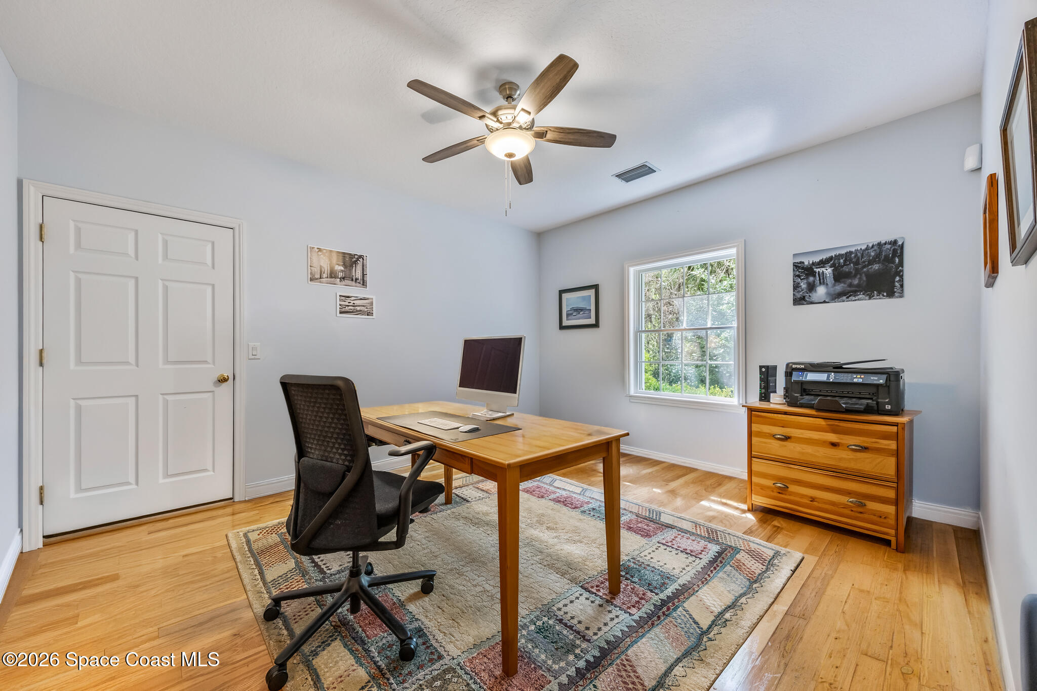 Home office with a wooden desk, patterned rug, hardwood floors, and a window overlooking greenery.