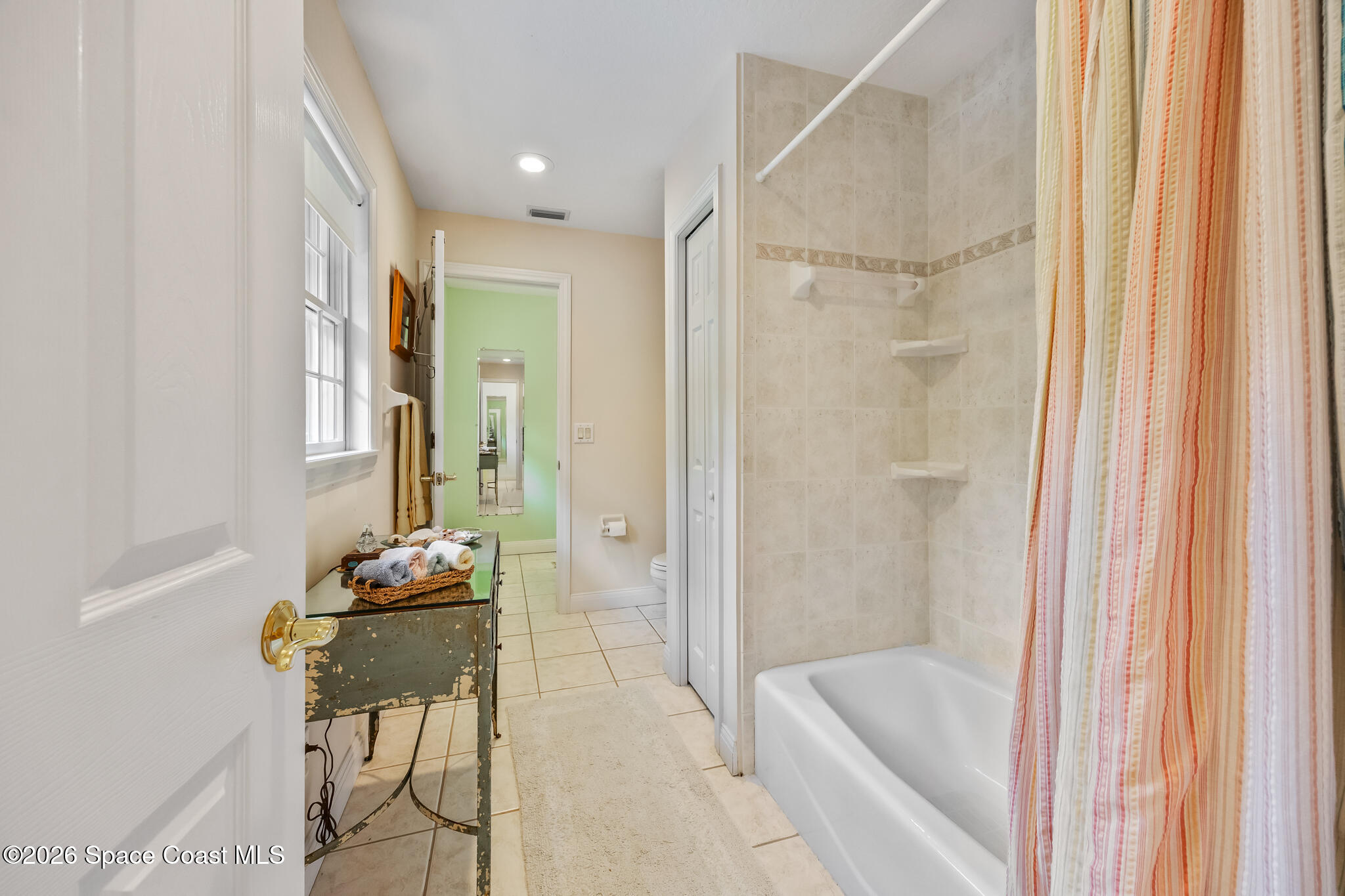 Bathroom with a tiled tub and shower, striped curtain, and distressed vanity table by a window.
