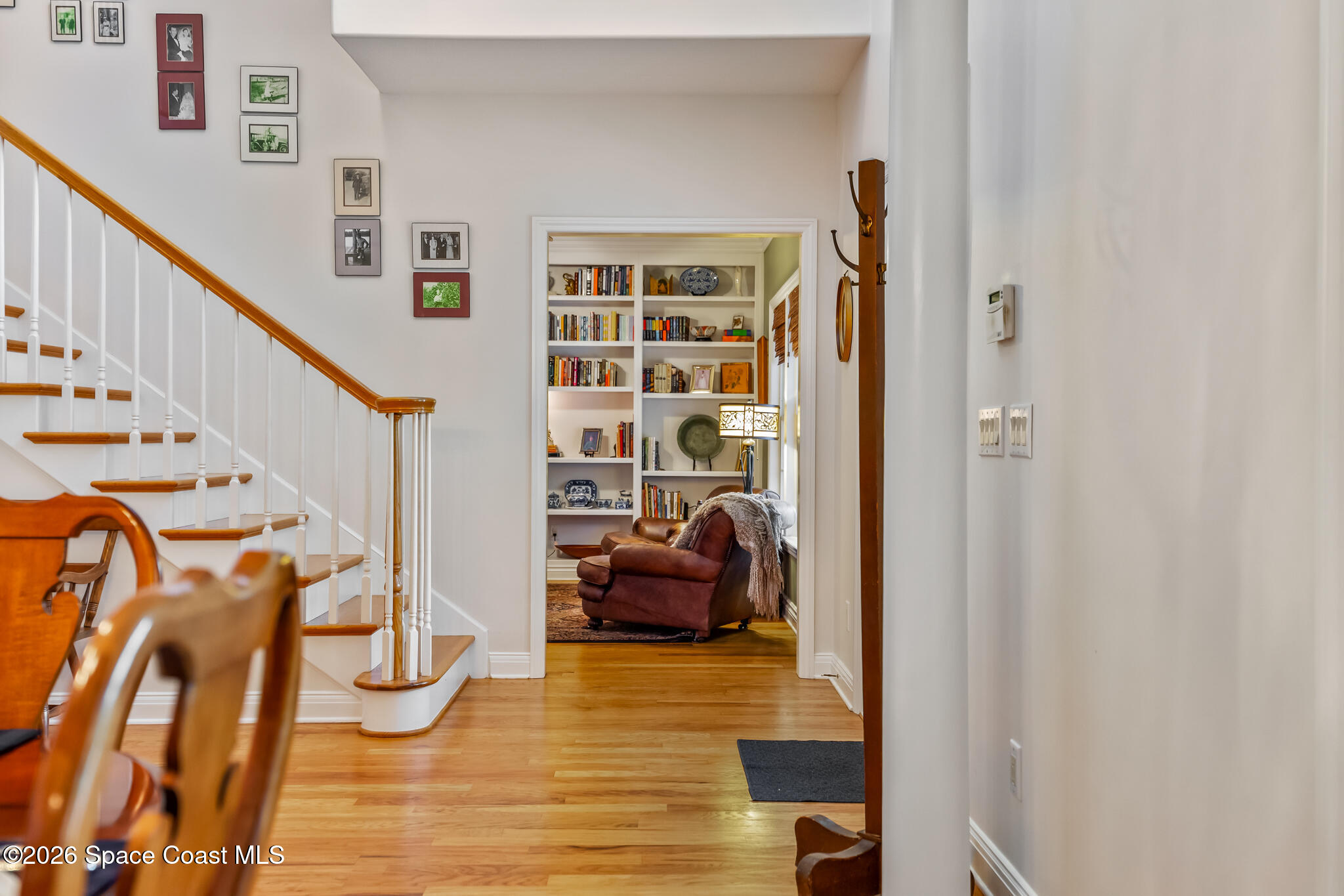Hardwood hallway beside a white staircase, looking into a sitting room with bookshelves and a leather armchair.