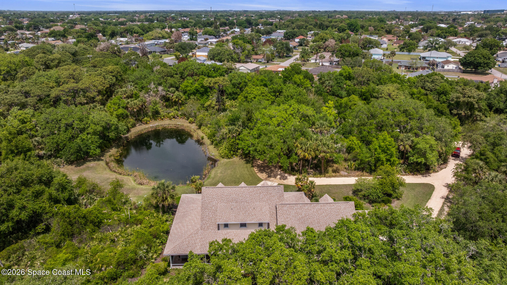 Aerial view of a wooded property with a pond, lawn, and home roof in the foreground.
