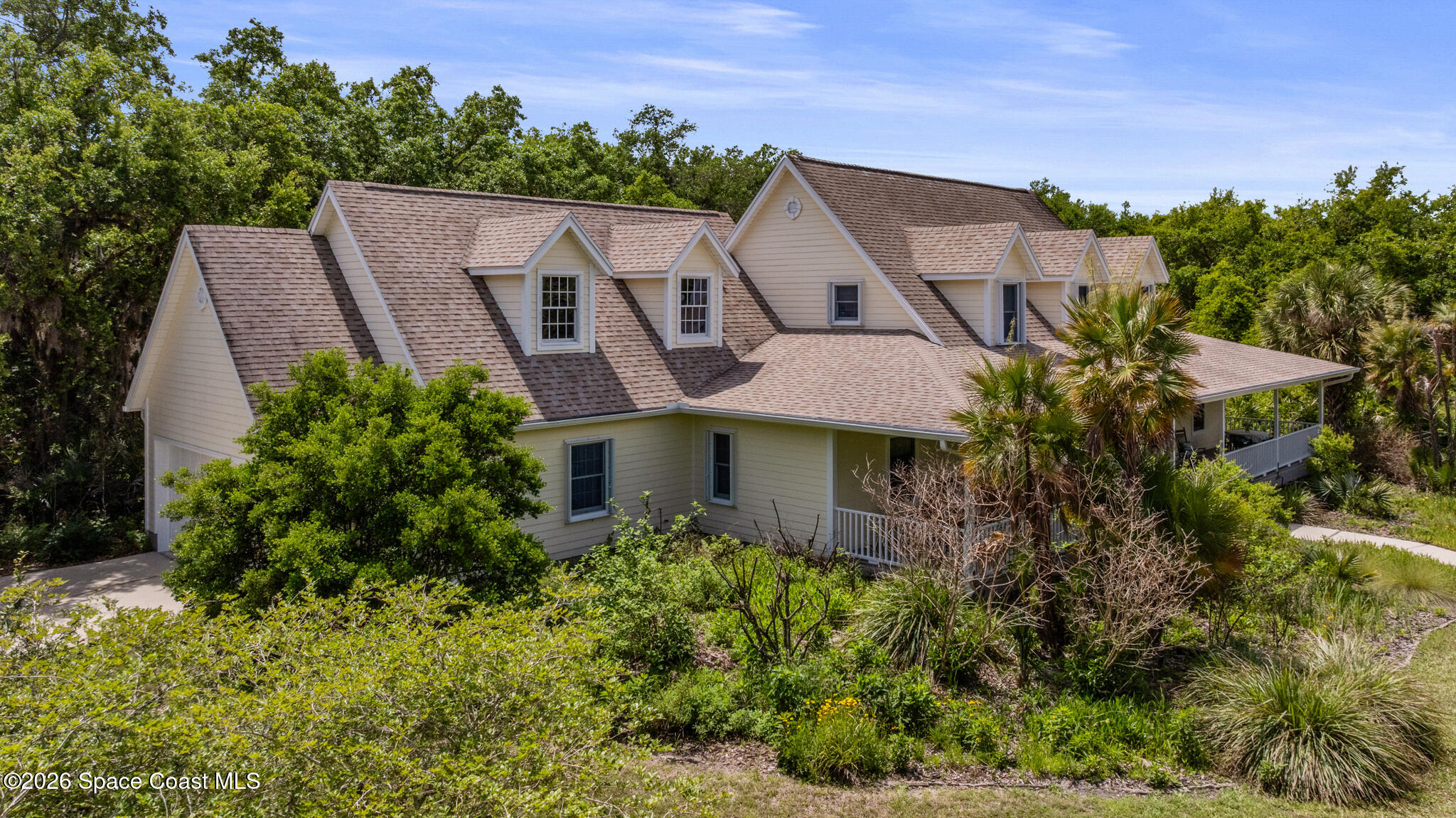 Beige multi-gabled home with a shingled roof, covered porch, and tropical landscaping.