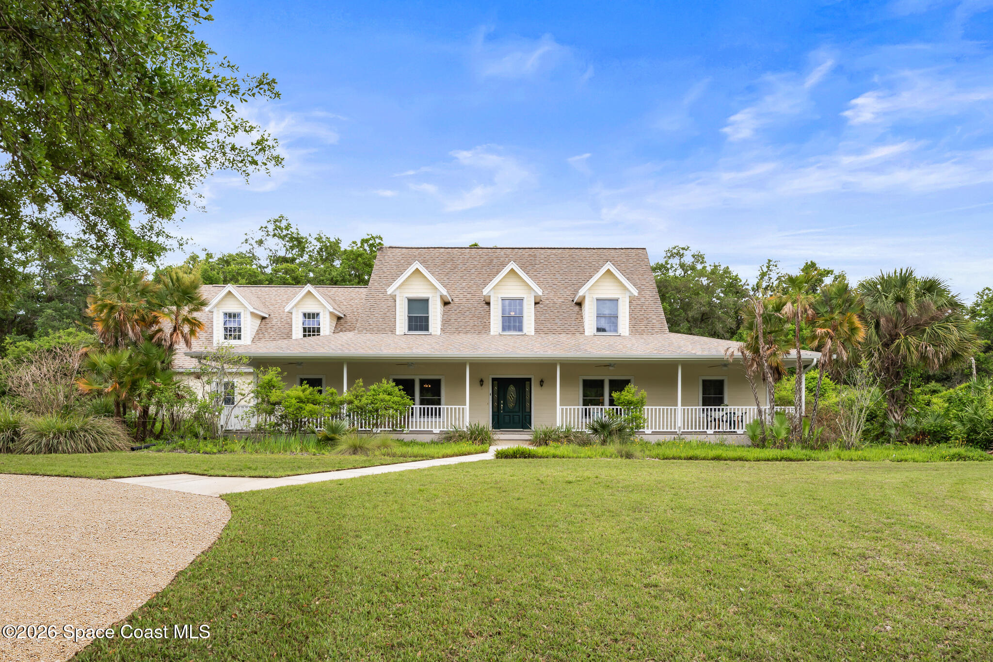 Cream two-story home with a wide covered porch, dark green door, dormers, palms, and a curved gravel driveway.