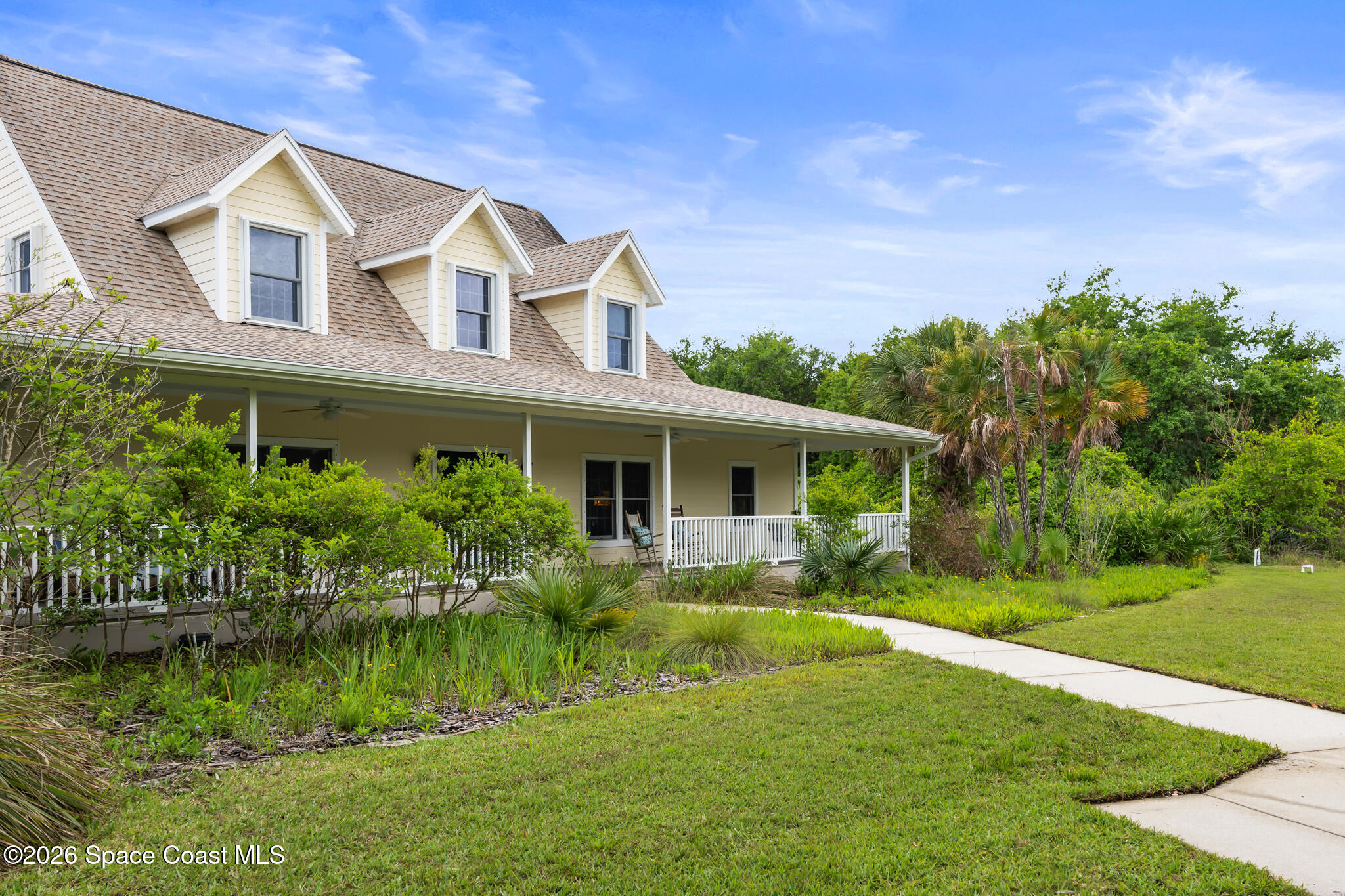 Two-story light-colored house with roof dormers, a covered porch, palms, and a curved walkway.