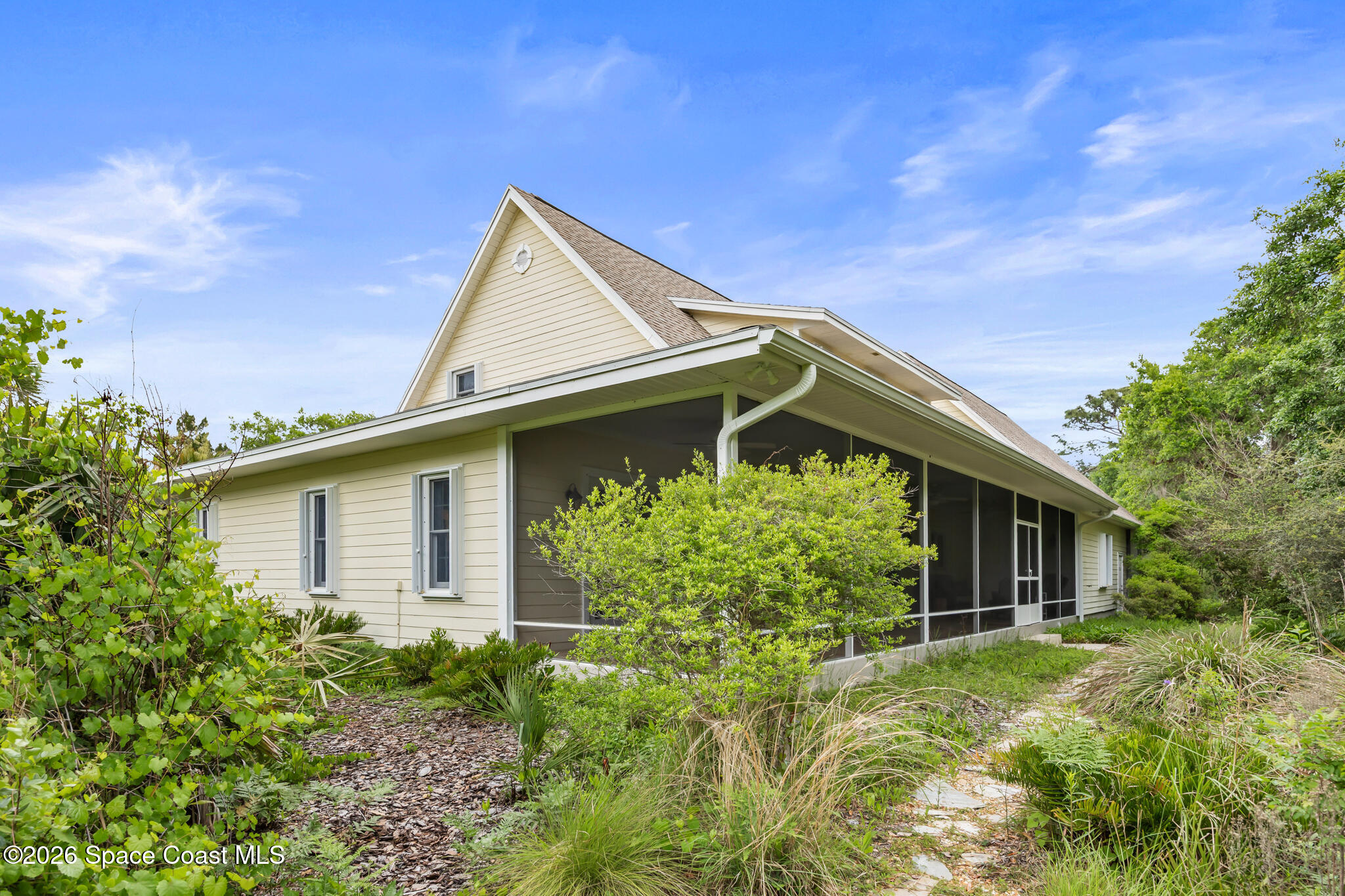 Cream-colored house with a steep gabled roof and screened porch amid lush landscaping.
