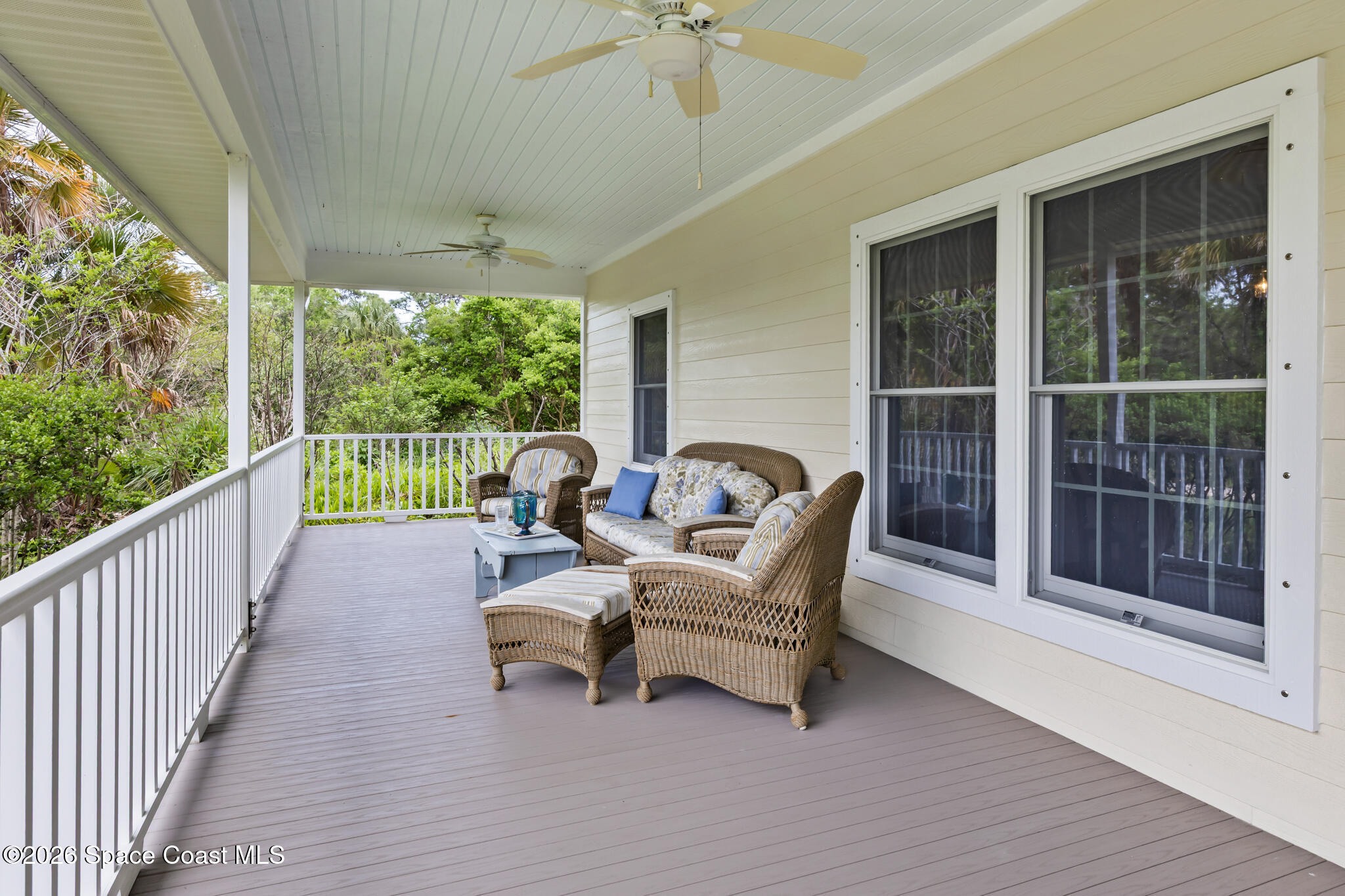 Covered porch with wicker seating, gray decking, white railings, and leafy trees beyond.