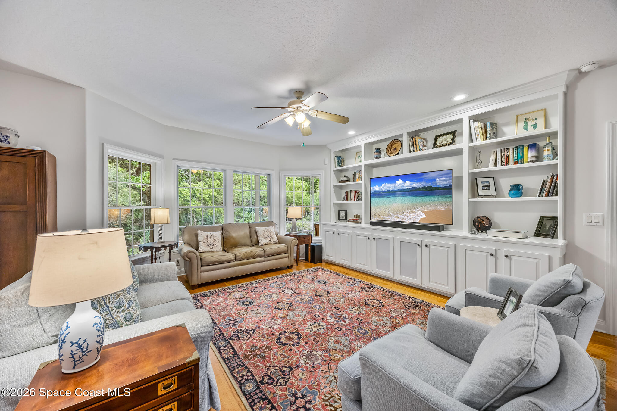Furnished living room with built-in white entertainment center, hardwood floors, and windows overlooking greenery.
