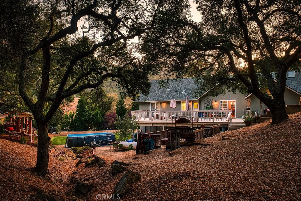 House with a raised deck and above-ground pool beneath large oak trees at dusk.
