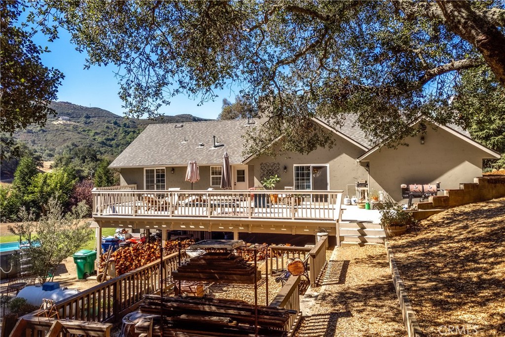 Rear of a tan home with a wide wooden deck and a partially visible pool, set against wooded hills.