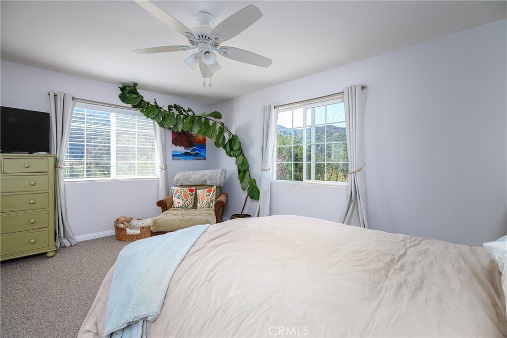 Bedroom with a large bed, ceiling fan, and windows overlooking tree-covered hills.