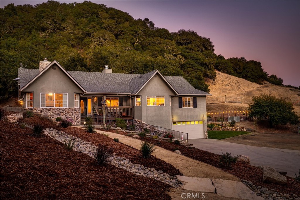 Gray hillside home with stone accents, a front porch, attached garage, and rock-lined garden path at dusk.