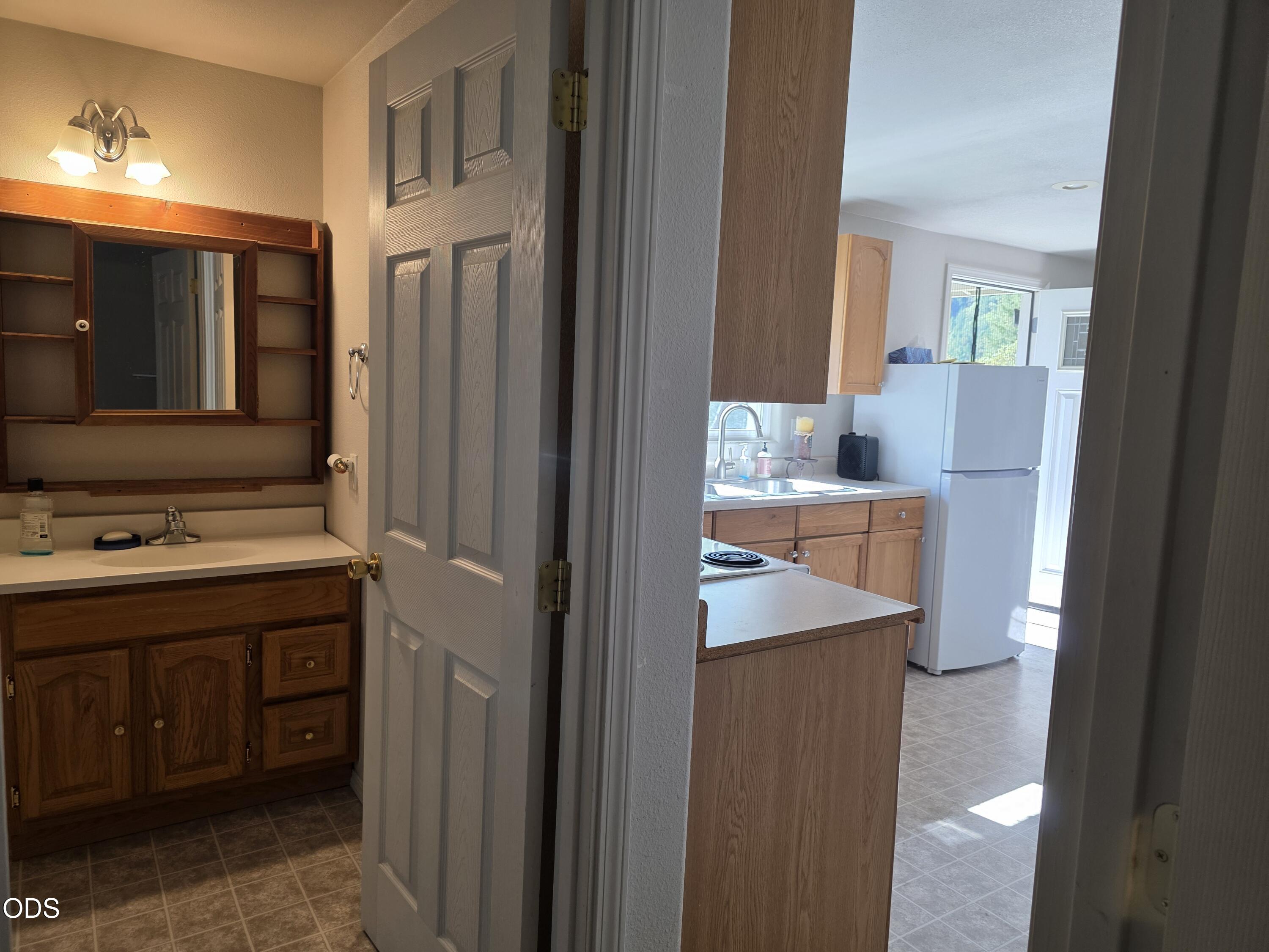 Bathroom vanity and mirror look toward a kitchen with wood cabinets and a white refrigerator.