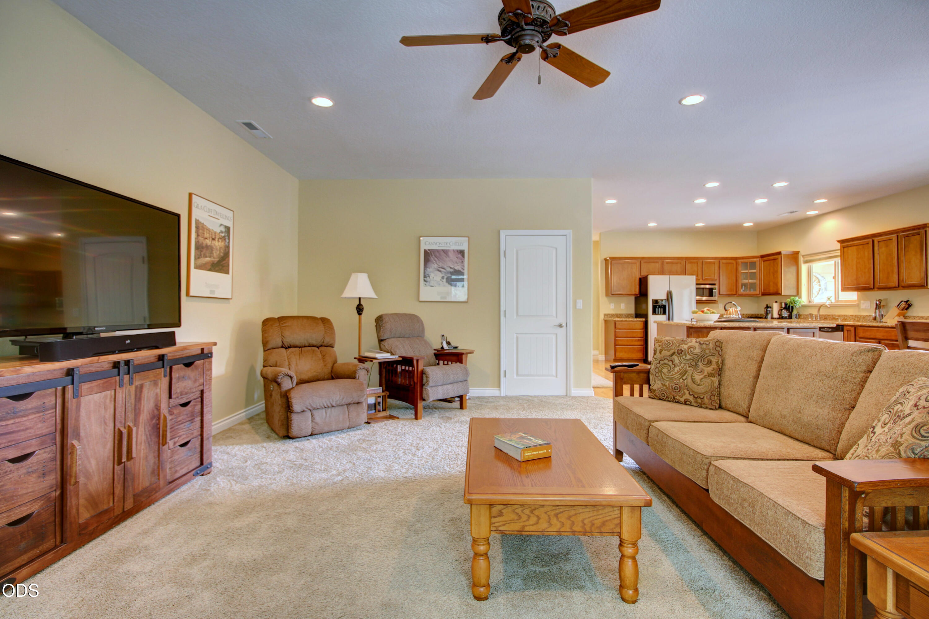 Furnished living room opening to a wood-cabinet kitchen, with beige carpet, recliners, and a ceiling fan.