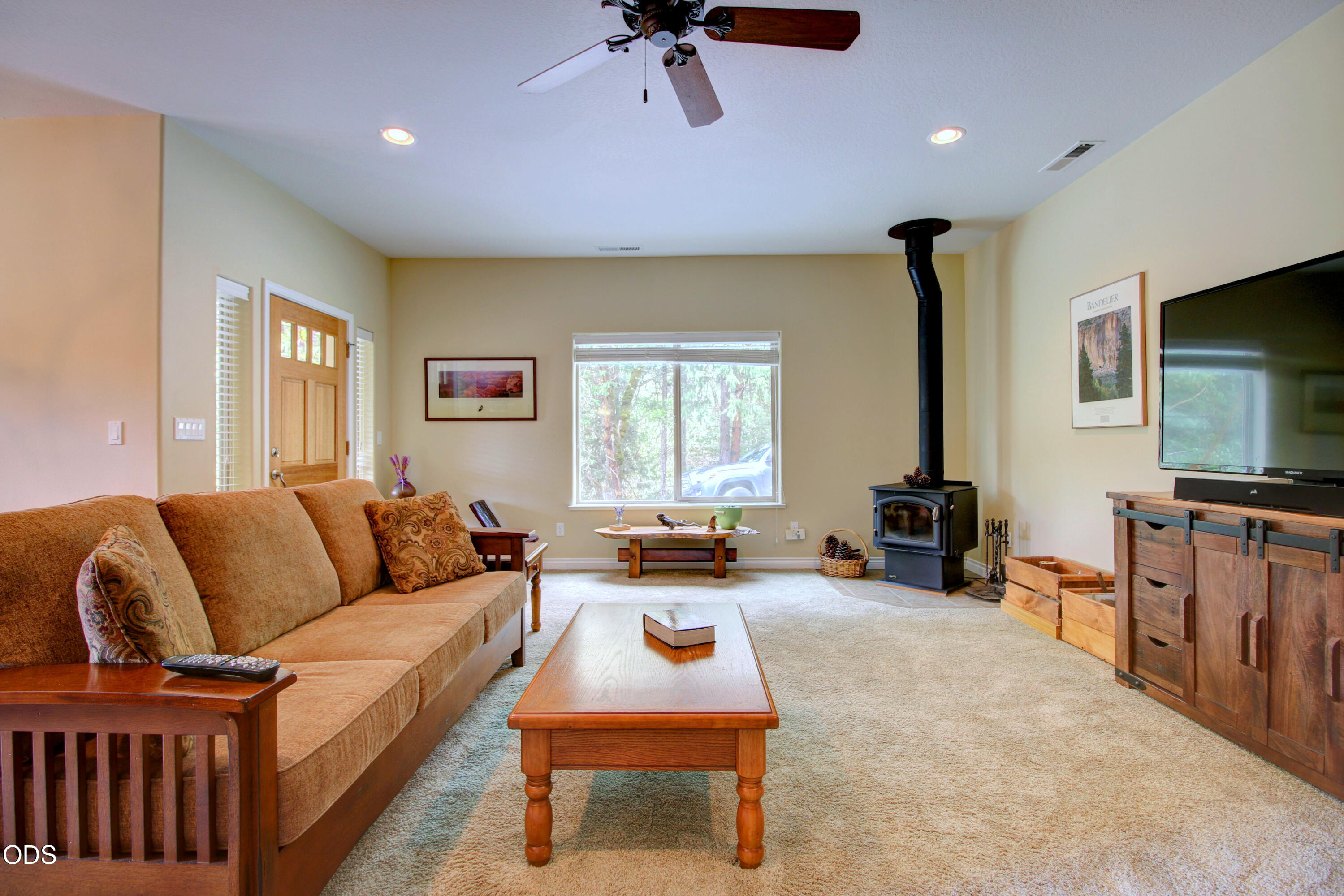 Living room with a tan sofa, wood-burning stove, and large window overlooking trees.
