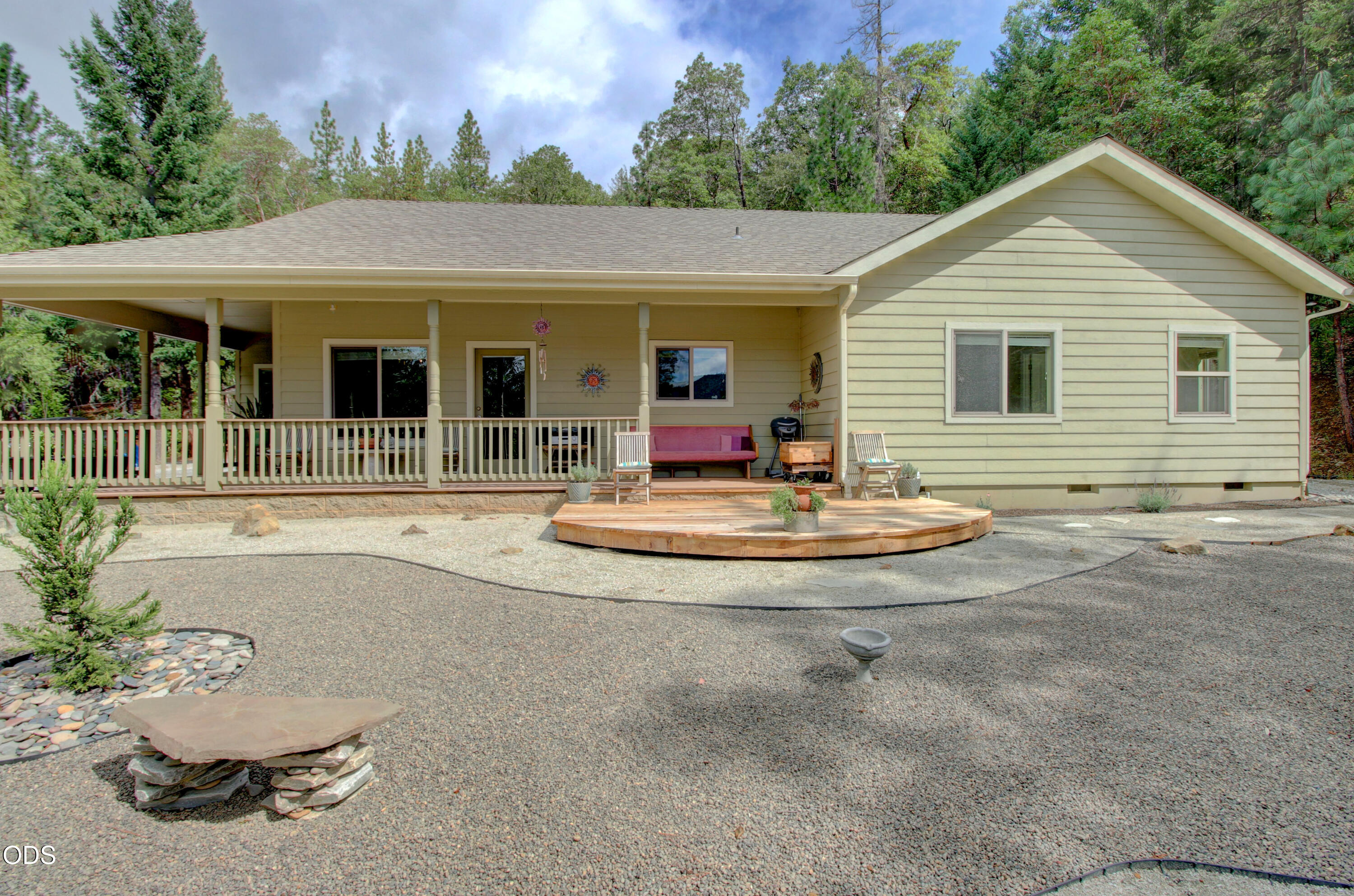 Home’s rear exterior with a covered porch, curved wooden deck, and gravel yard among trees.