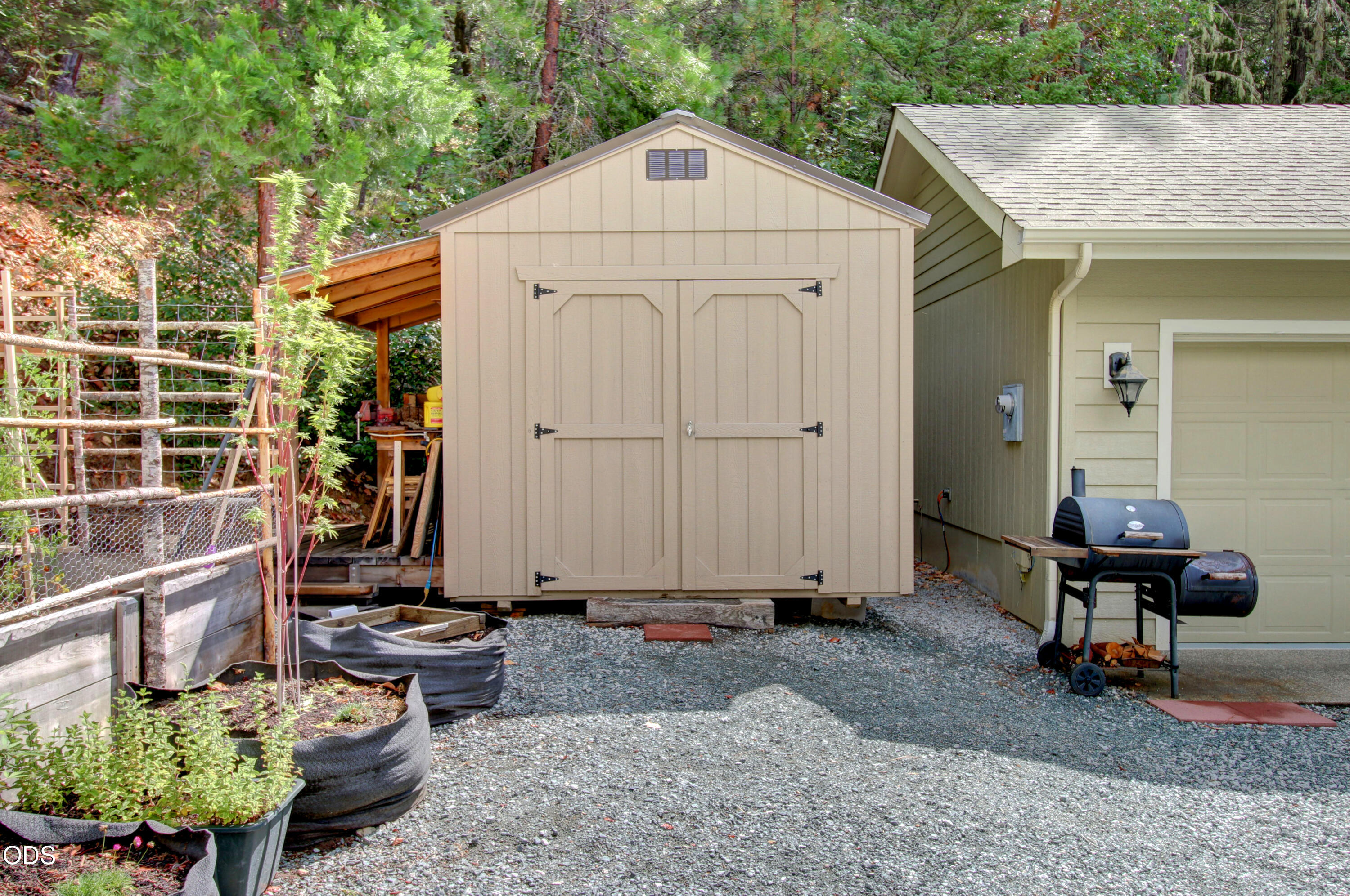 Beige storage shed beside a garage, with raised garden beds and a barbecue on gravel.