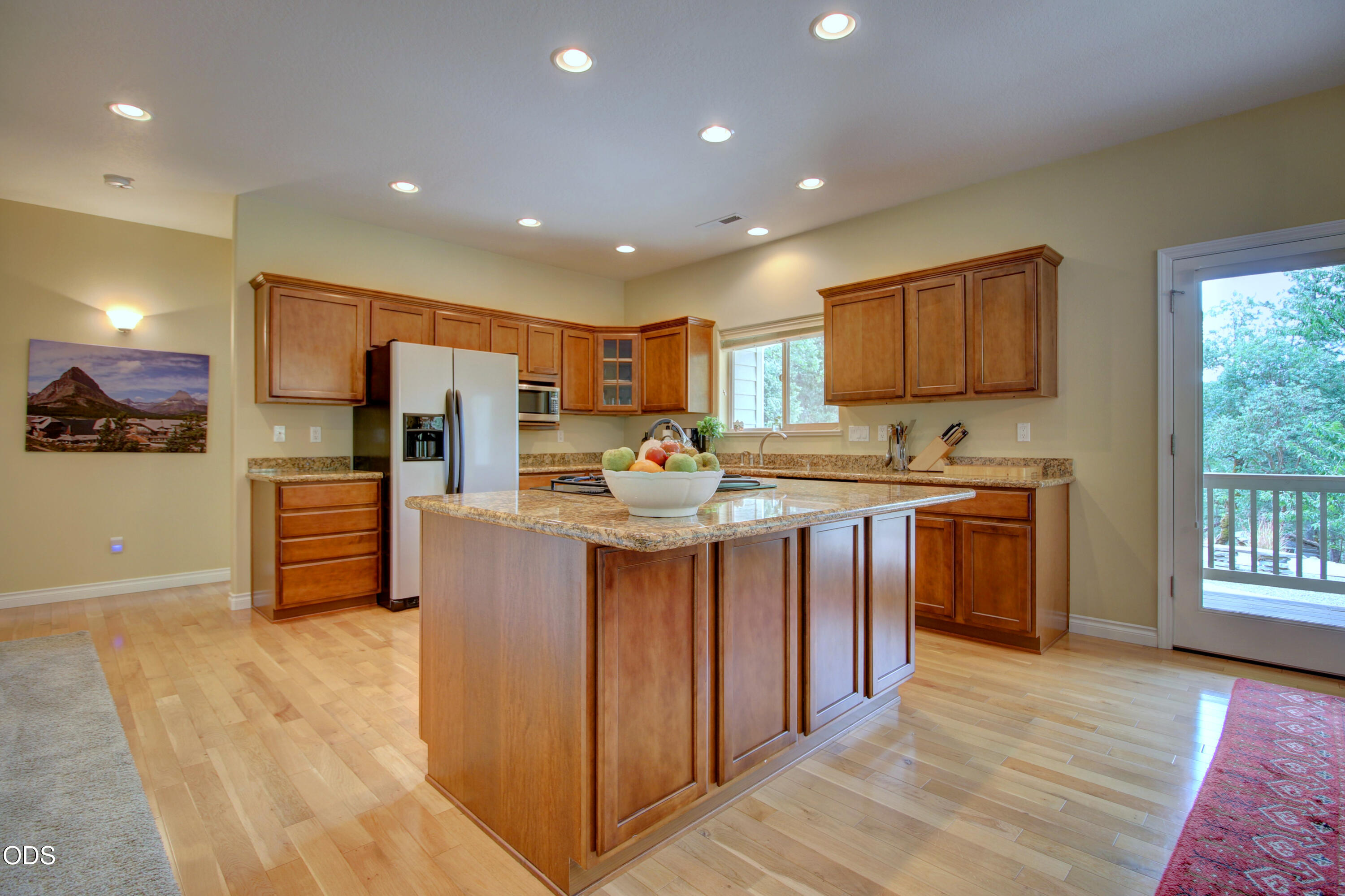 Kitchen with a large granite-topped island, warm wood cabinets, and a glass door opening to a leafy view.