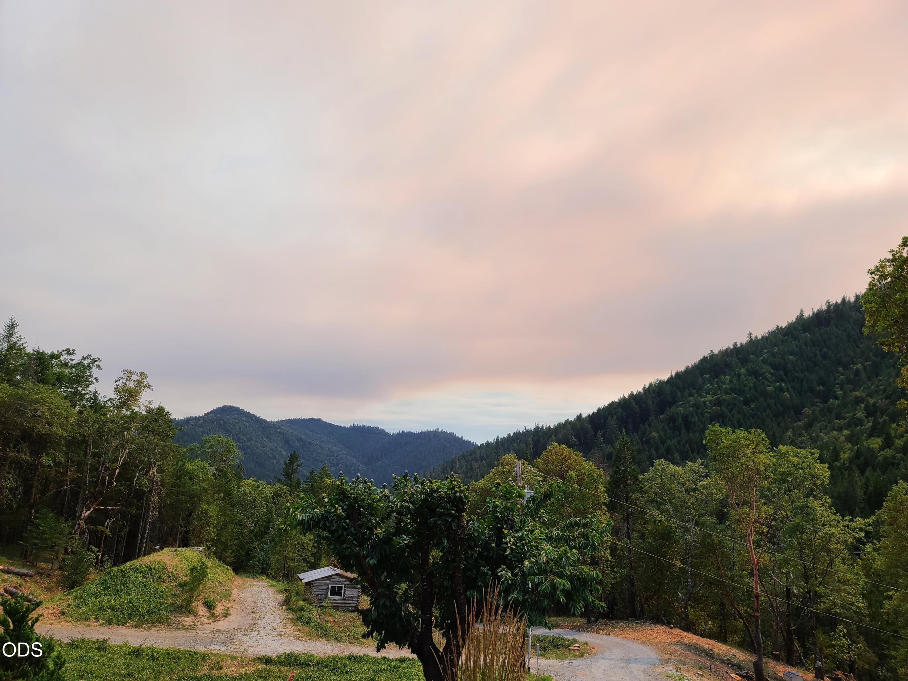 Wooded mountain ridges beyond a gravel drive and small shed under a pink evening sky.