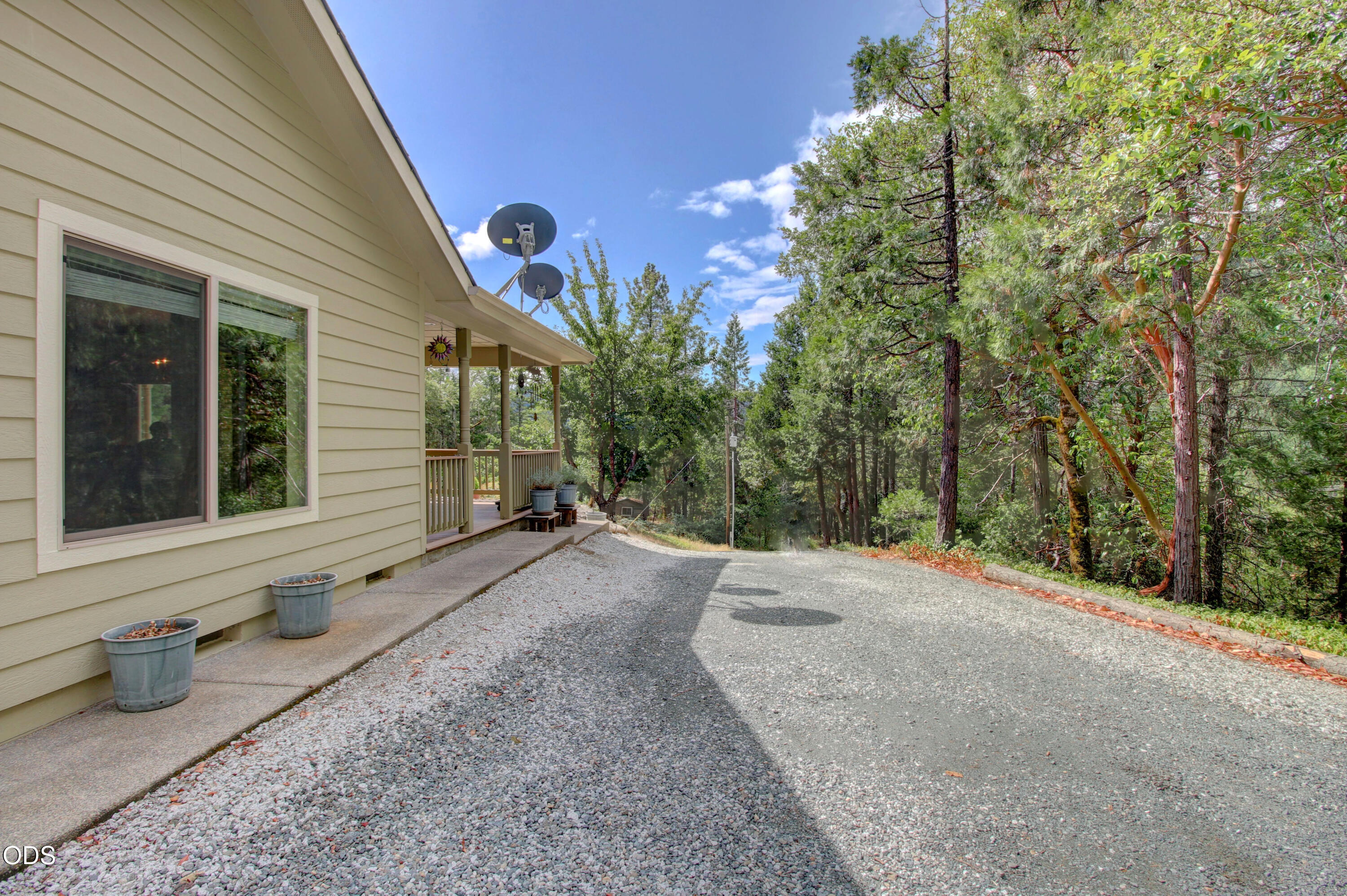Beige-sided house and covered porch beside a gravel driveway in a wooded setting.