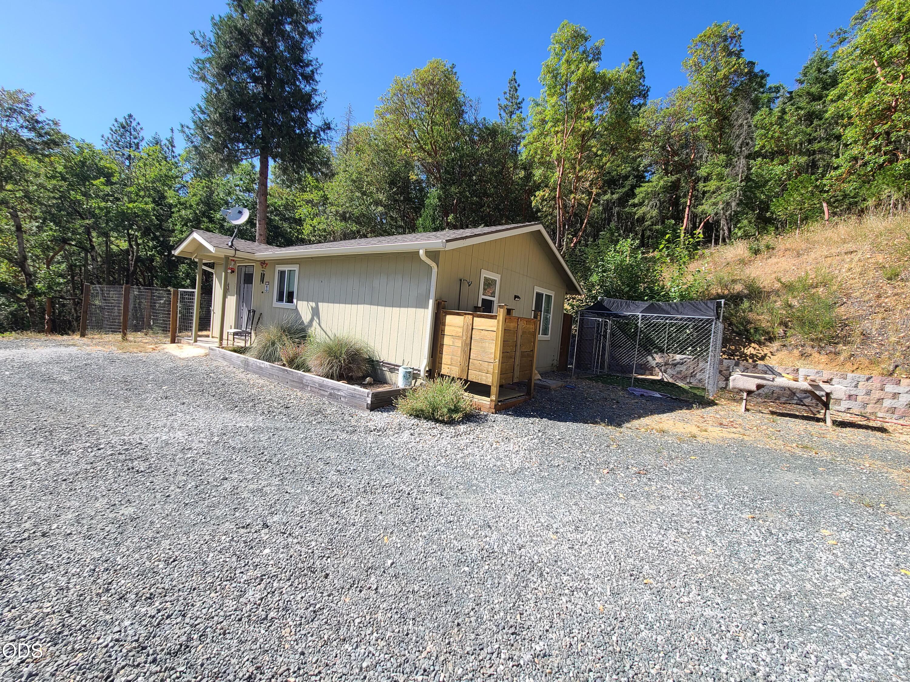 Small single-story home beside a gravel driveway, with a wooden entry platform and fenced enclosure.