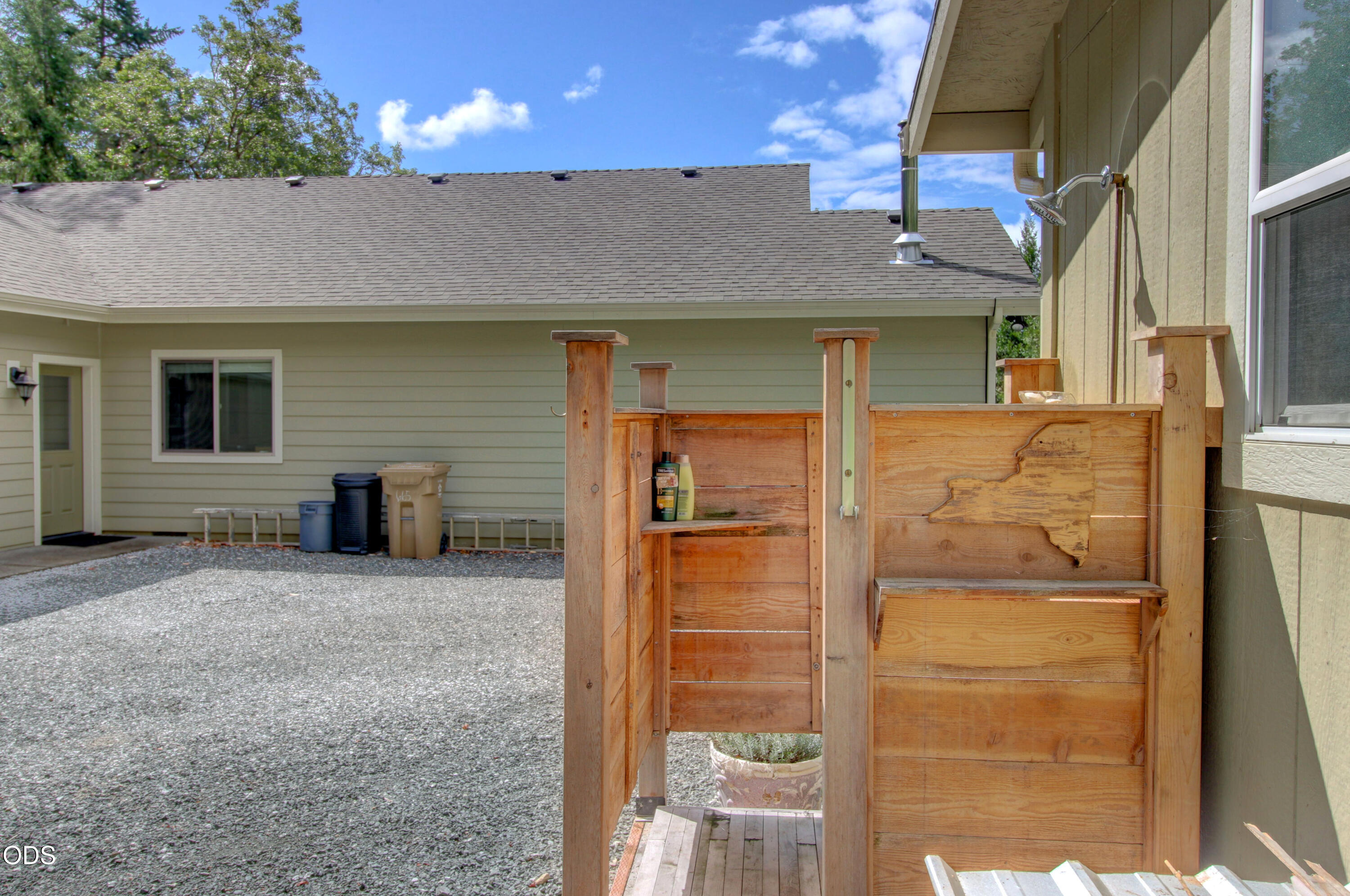 Wooden outdoor shower enclosure beside a house, with a wall-mounted showerhead and gravel yard.