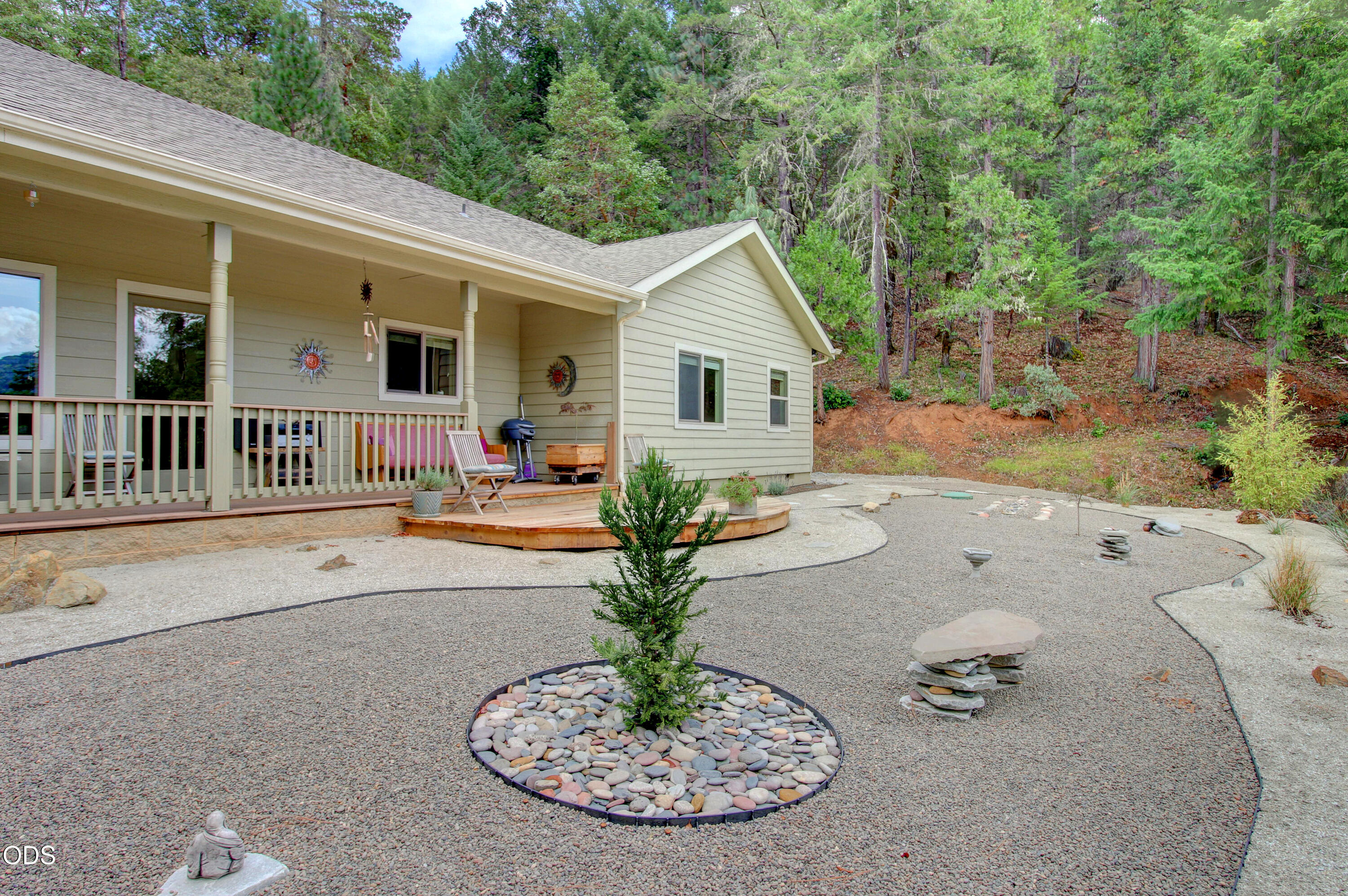 House with a covered porch and deck beside a gravel yard, backed by a wooded hillside.