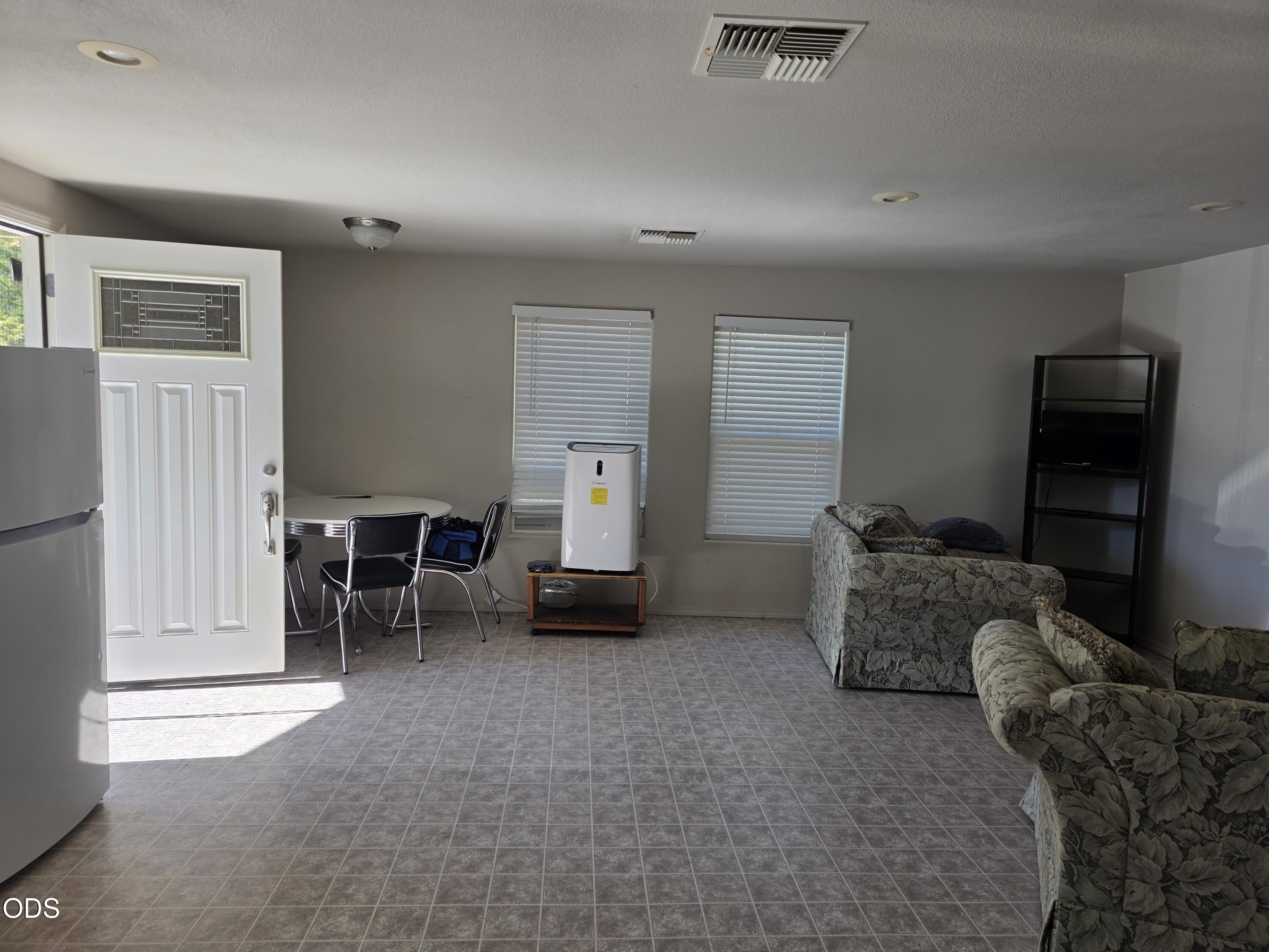Living and dining area with patterned sofas, tiled floors, and a portable air conditioner between two windows.