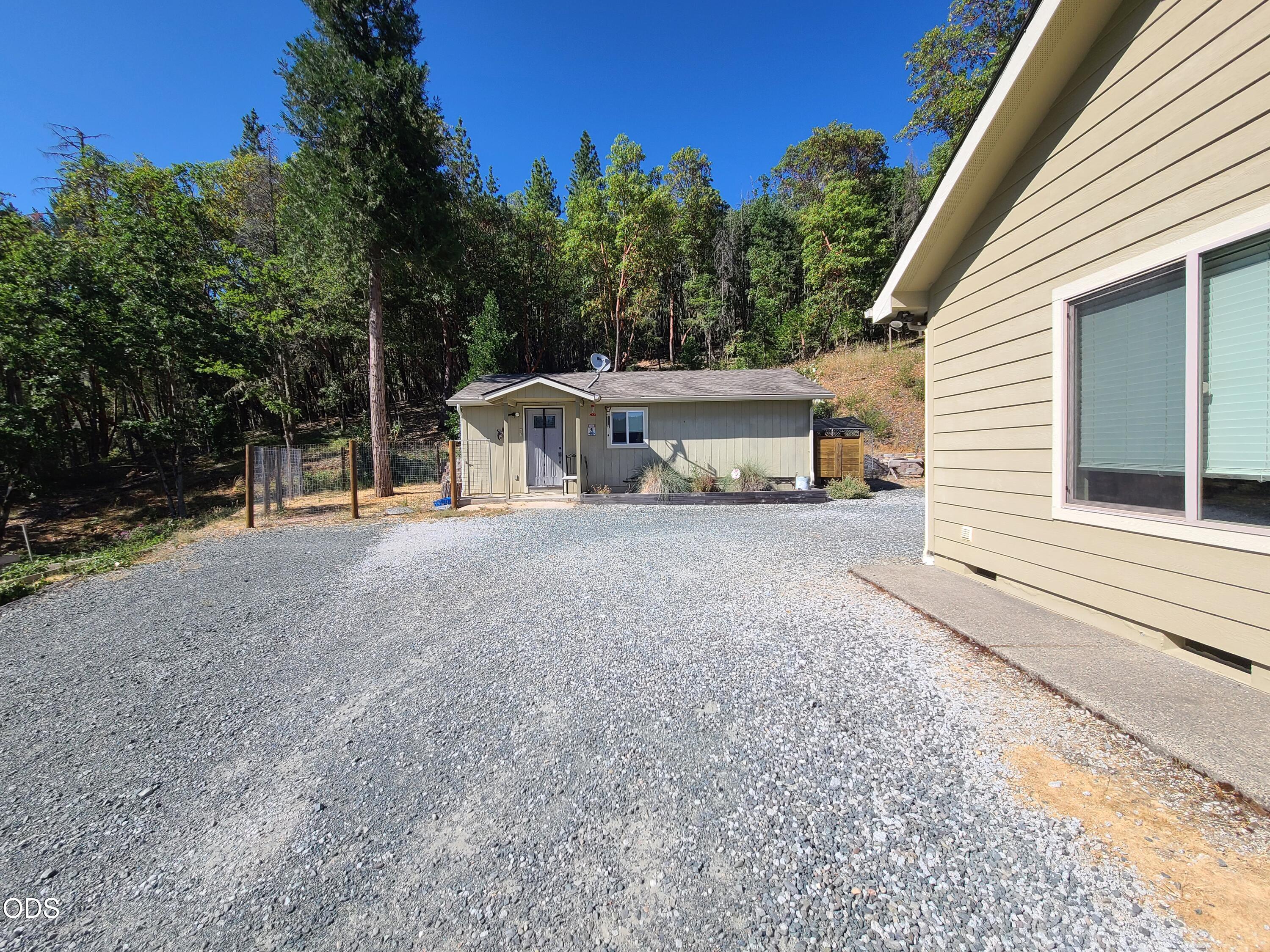Property exterior with a broad gravel driveway, detached building, and wooded hillside.