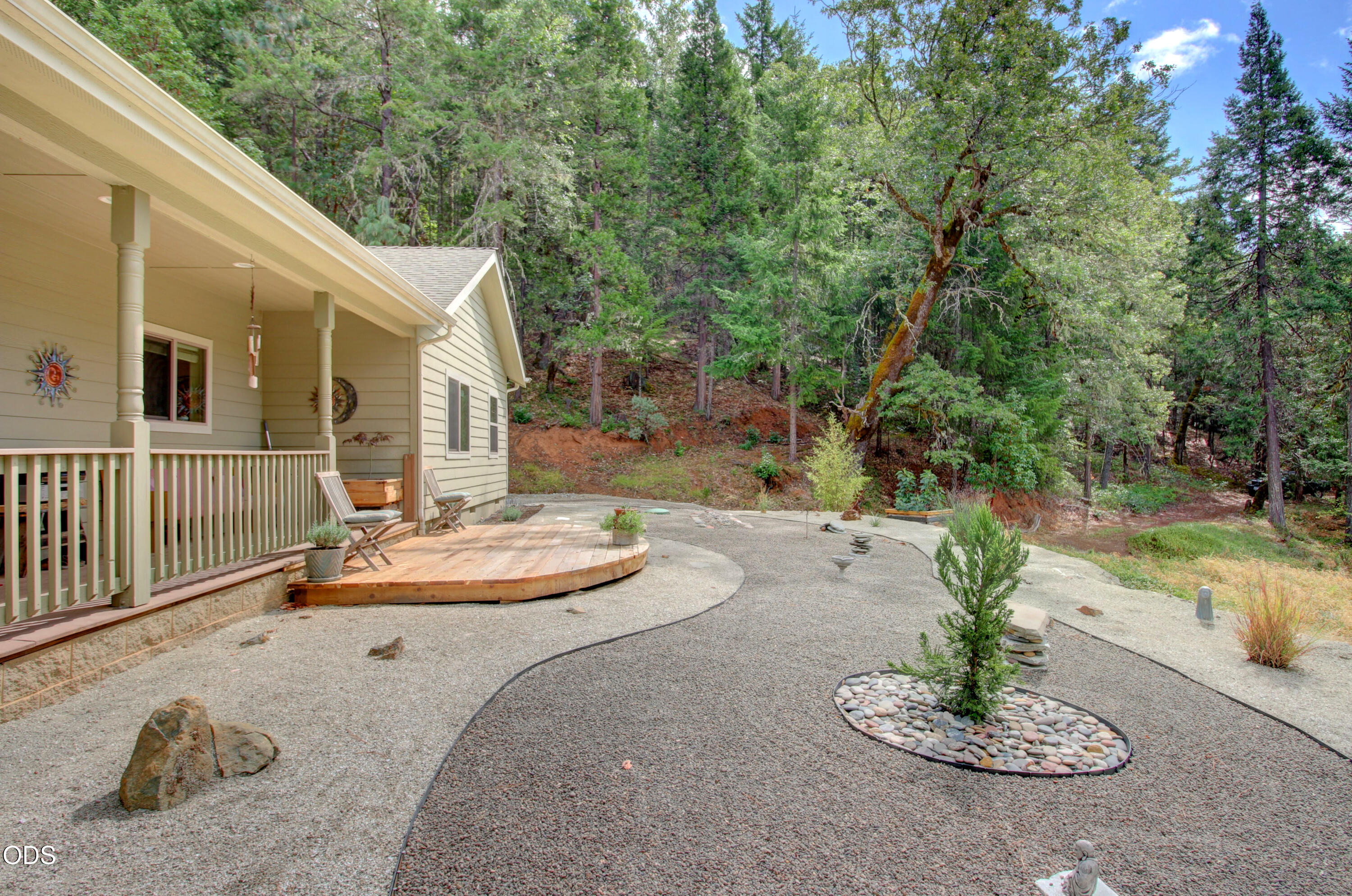 Home with a covered porch and wooden deck beside a landscaped gravel patio and wooded hillside.