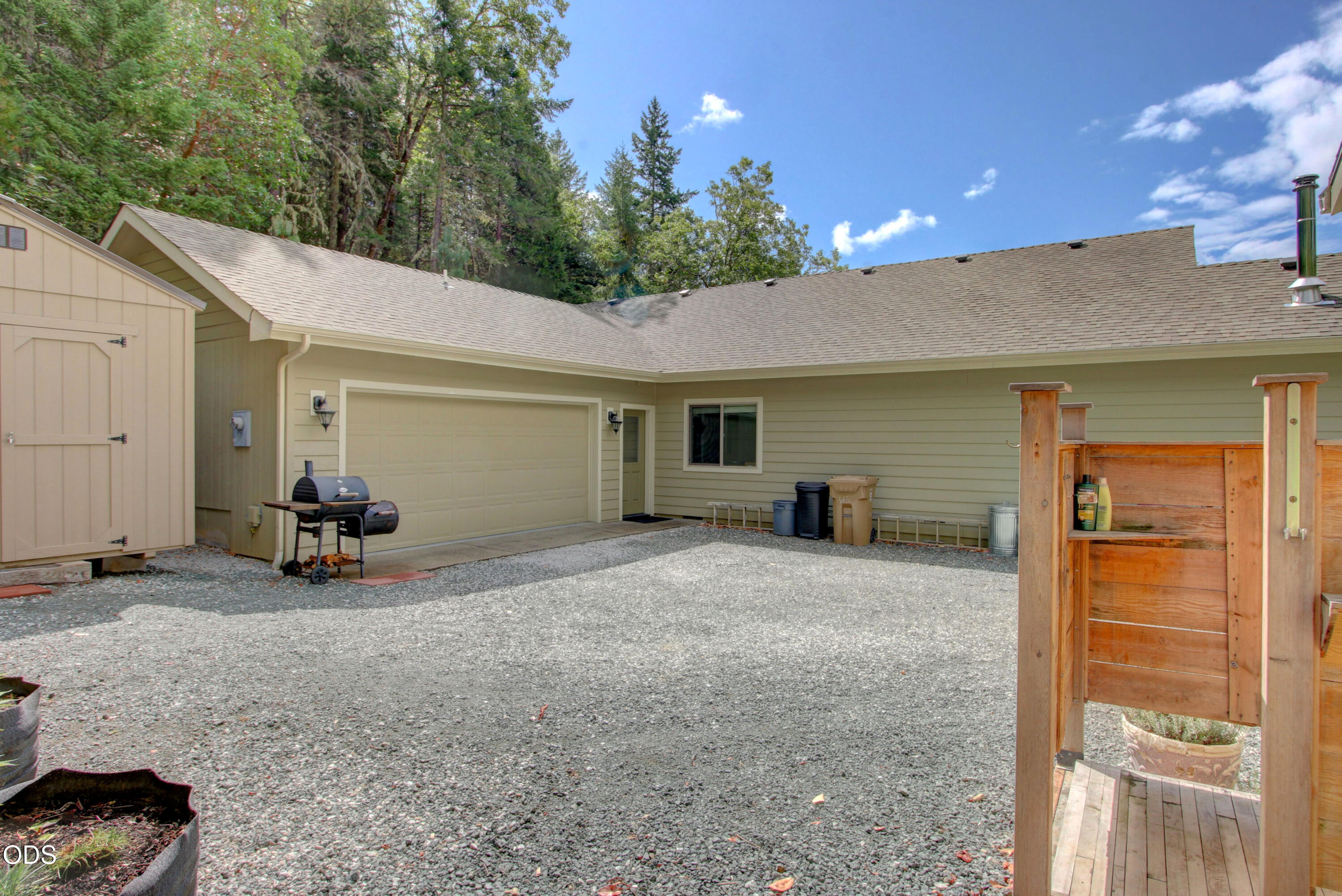 Home exterior with a two-car garage, gravel courtyard, and wooded backdrop.