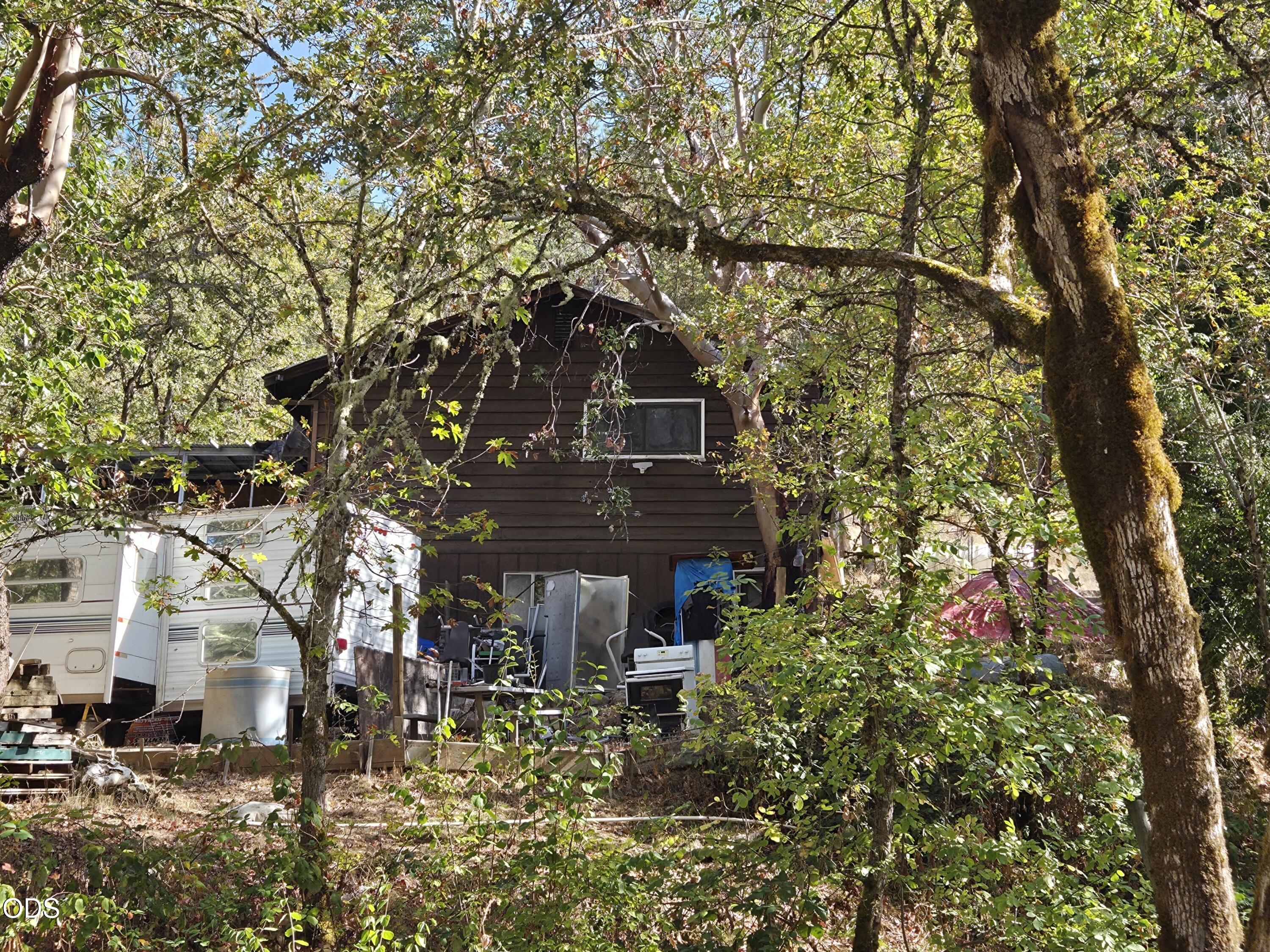 Wood-sided house tucked among dense trees, with a white travel trailer beside it.